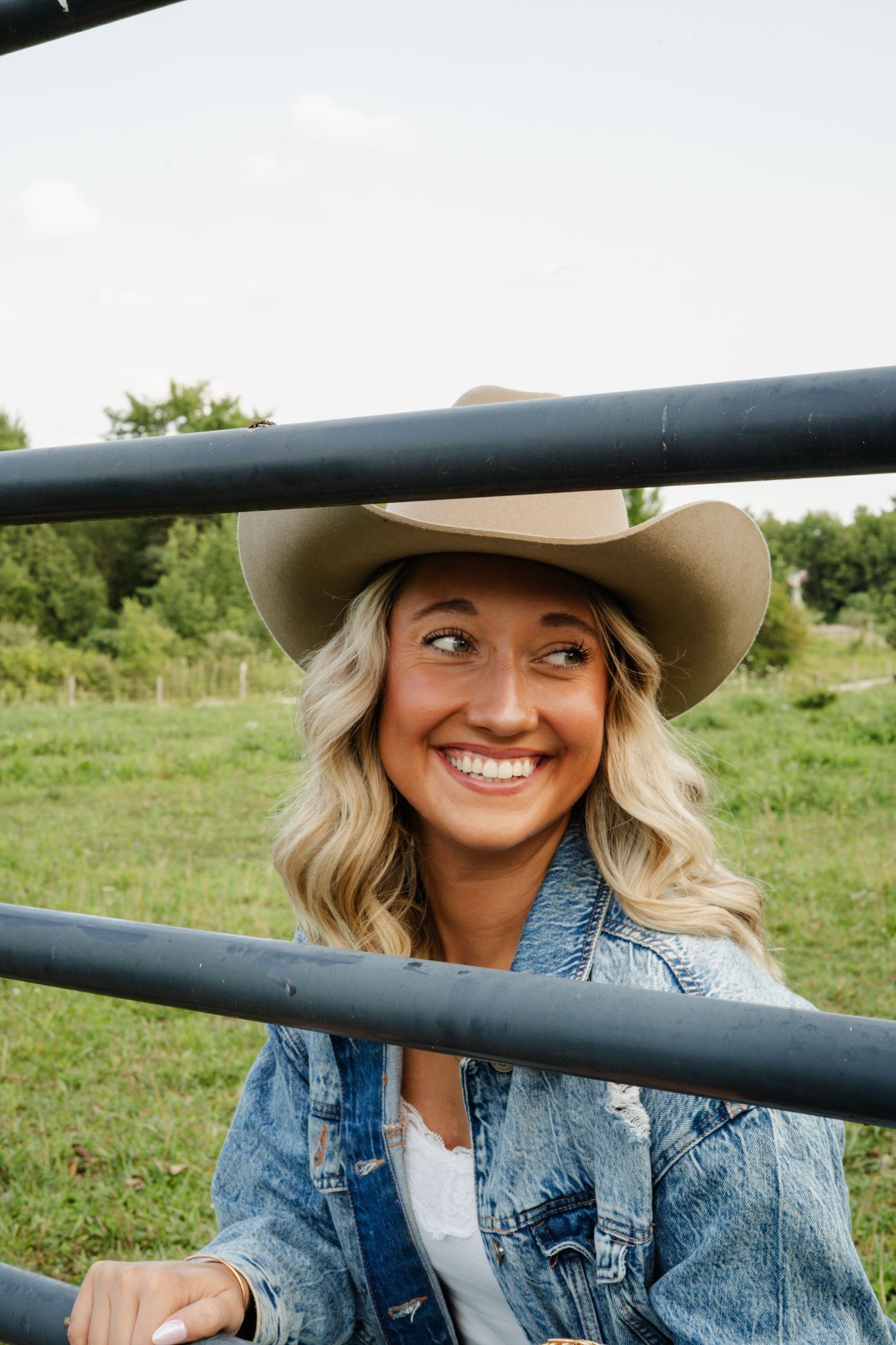Happy blonde woman in cowboy hat and denim jacket smiling through metal farm gate rails.