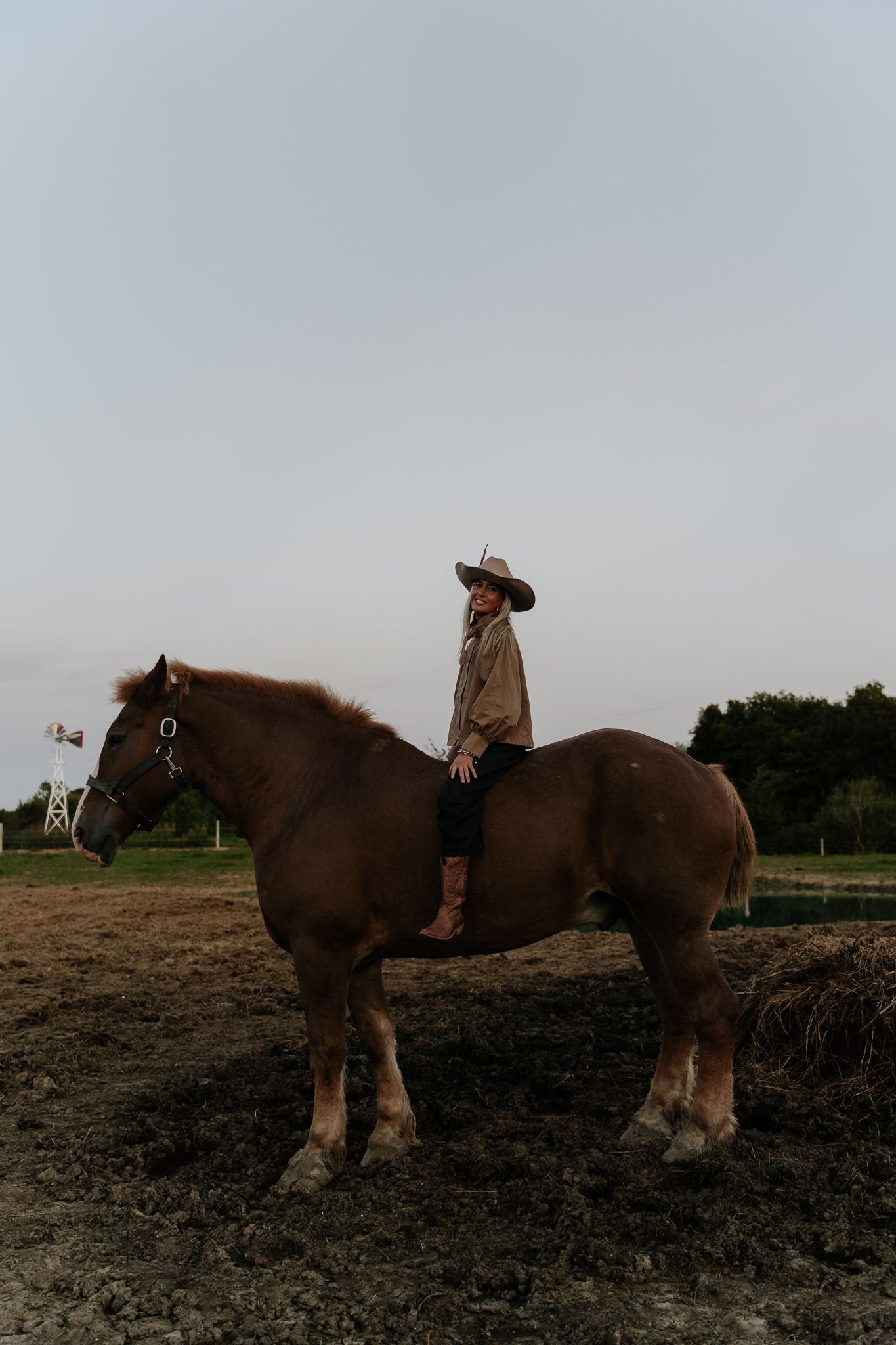 Woman in cowboy hat riding a large brown horse on a rural farm at dusk.