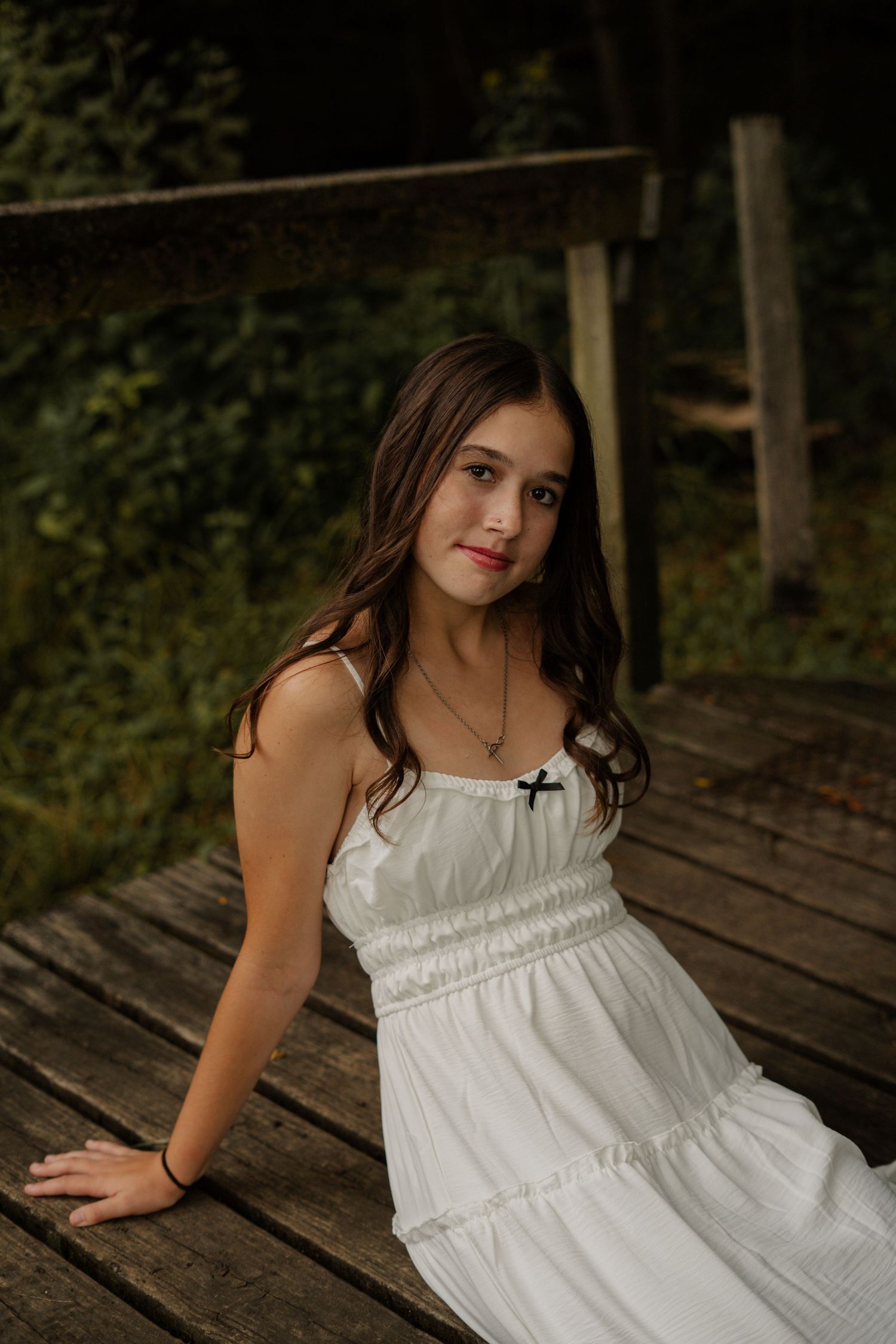 Young brunette woman in white sundress with cross necklace sitting on rustic wooden deck.