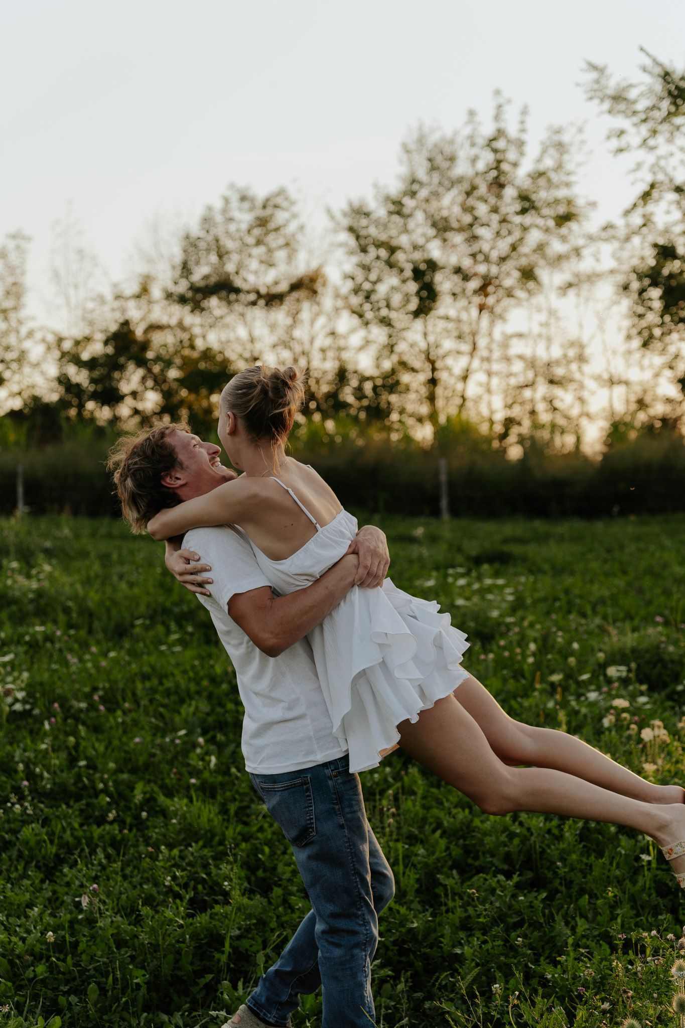 Man lifting woman in white dress in a sunlit green meadow at golden hour.