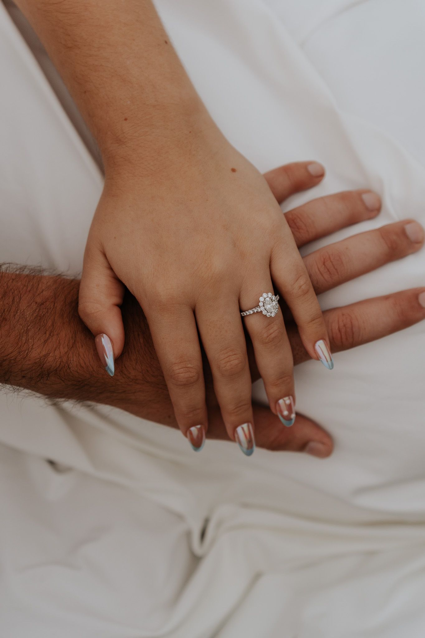 Close-up of engaged couple holding hands showing beautiful halo diamond engagement ring with white manicured nails.
