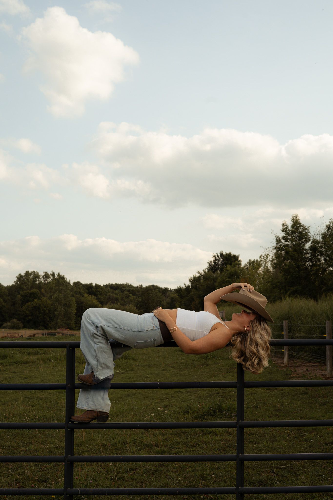 Blonde woman in cowboy hat and jeans posing on metal farm fence against cloudy sky background.