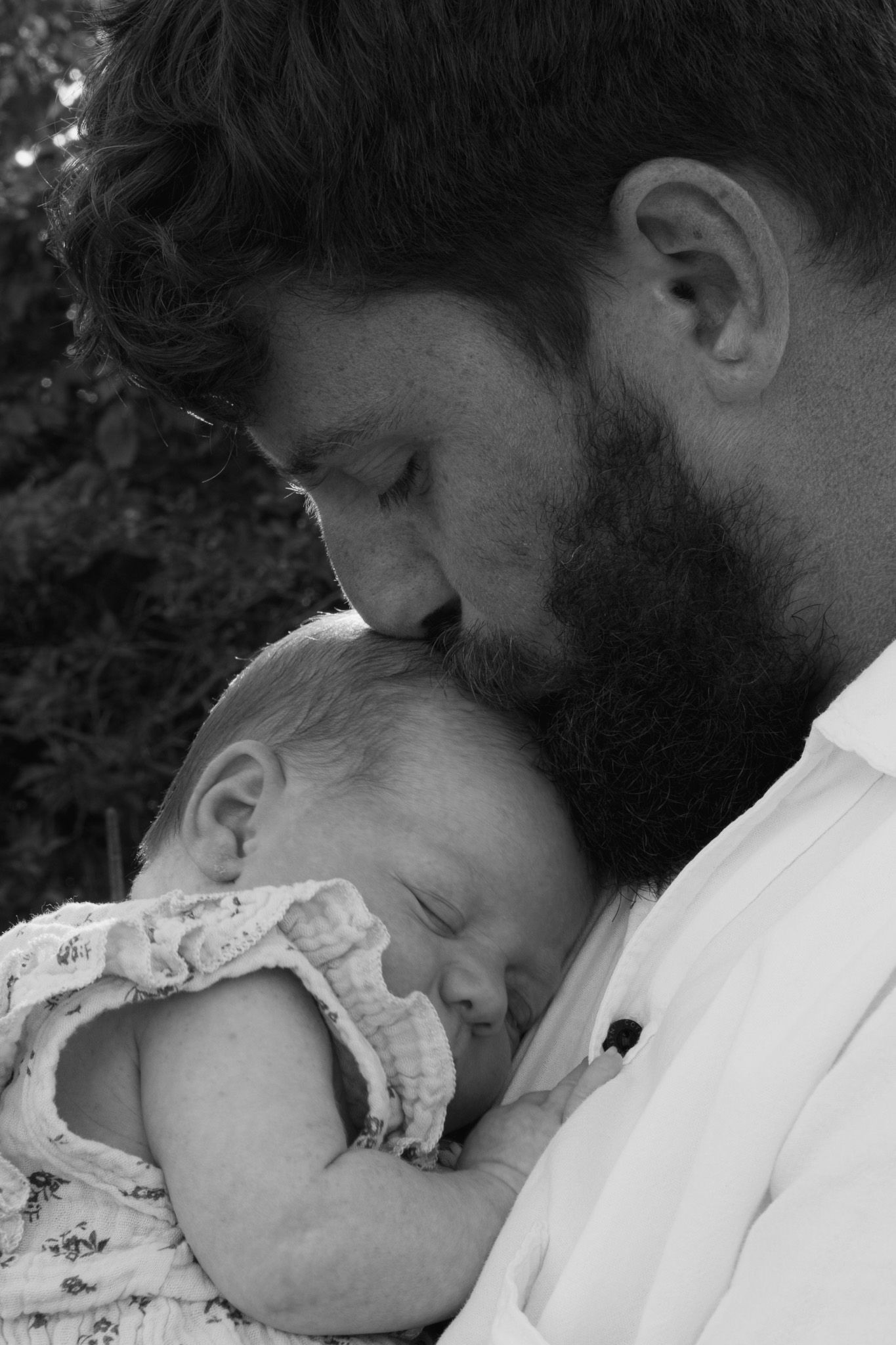 Bearded father tenderly kissing sleeping newborn baby in black and white portrait.