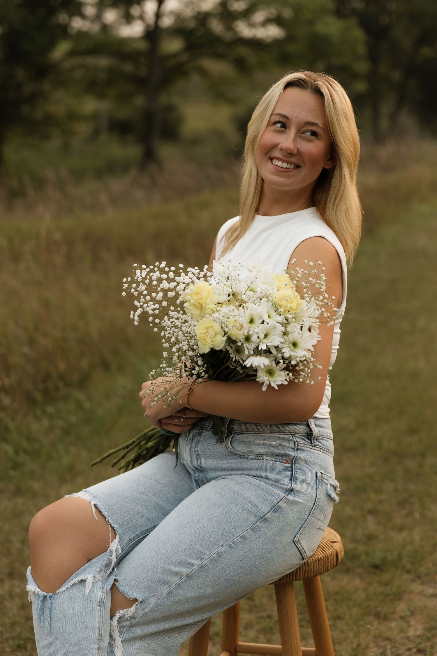 Smiling blonde woman sitting on wooden stool holding white flower bouquet wearing ripped jeans and white top outdoors.