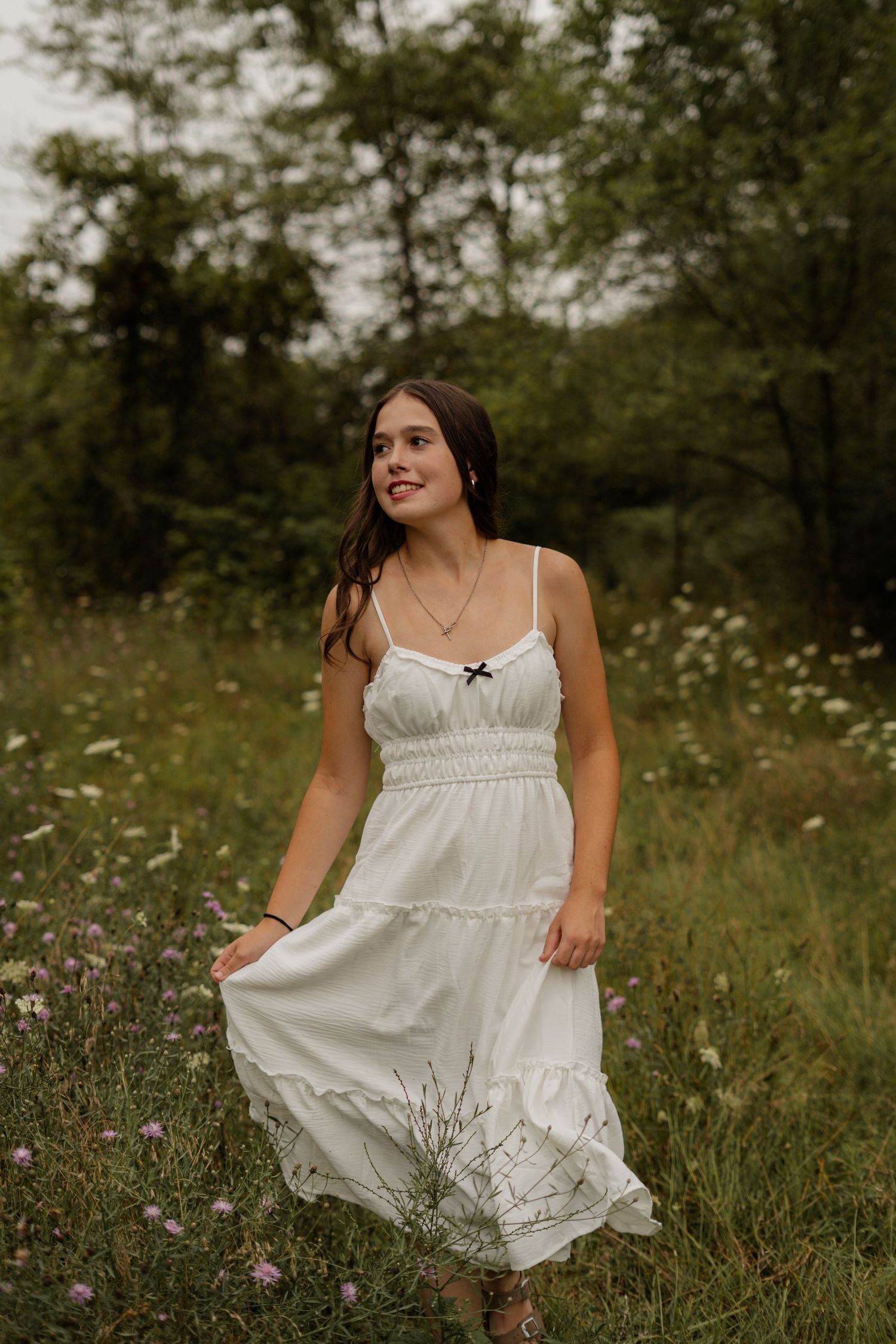 Young woman in white sundress walking through wildflower meadow with green forest background.
