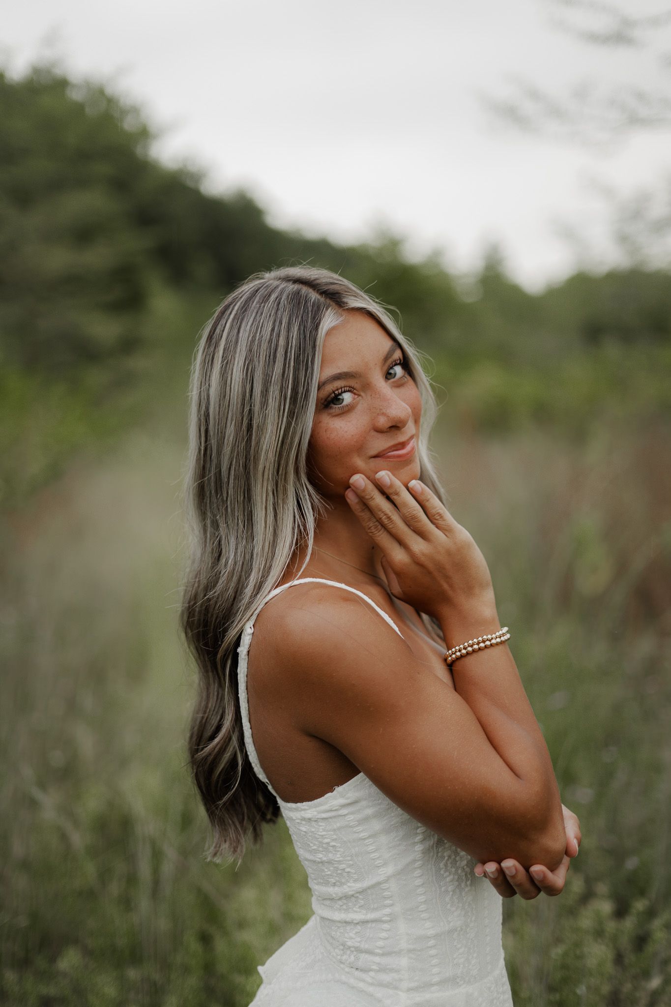 Young woman in white dress touching her face gently while posing in a green outdoor setting.