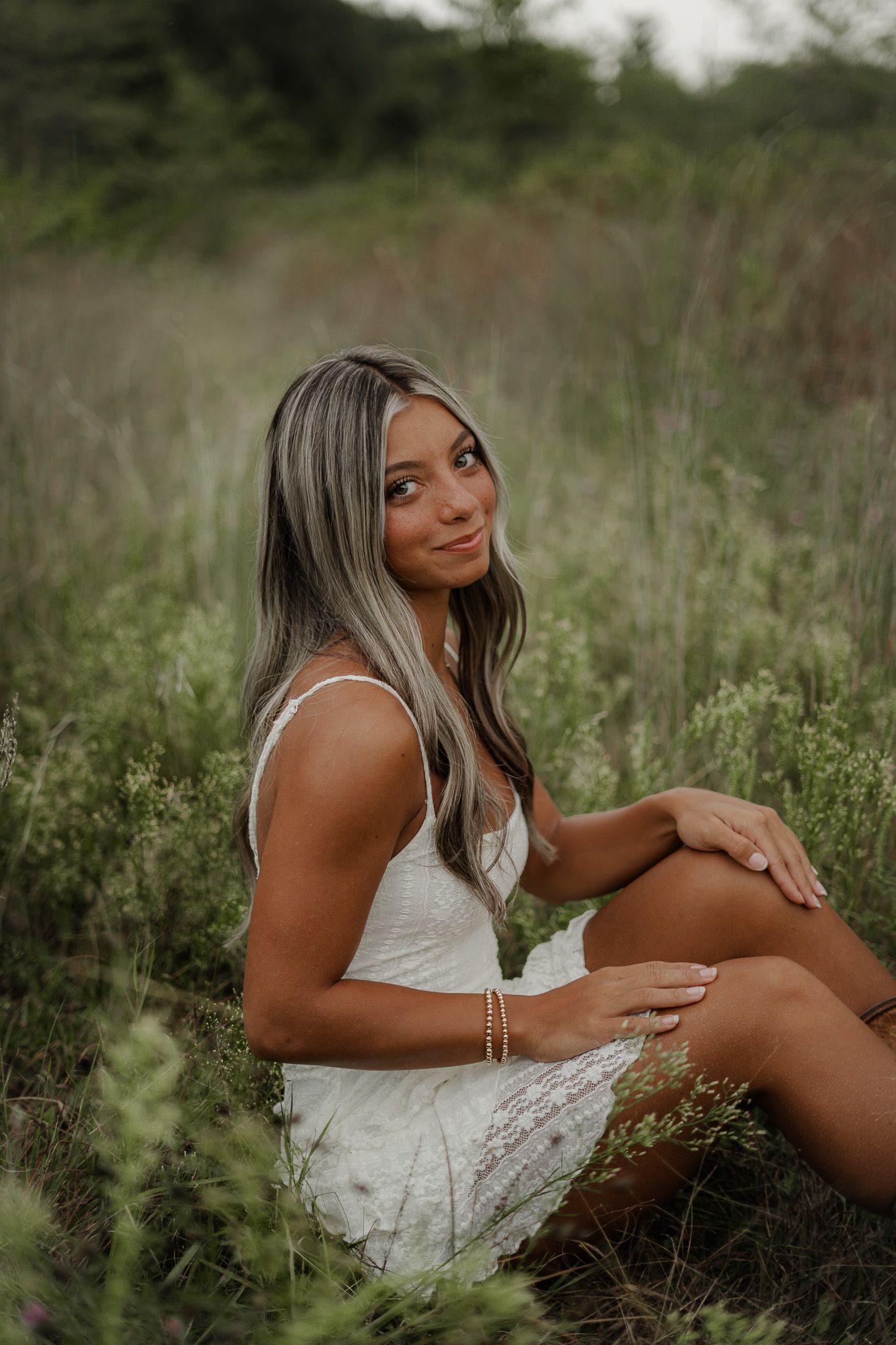 Young woman with highlighted hair in white dress sitting in lush green meadow for senior photos.