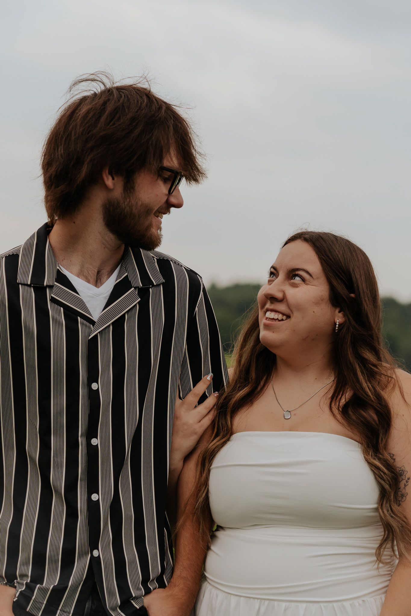 Happy engaged couple smiling at each other outdoors, woman in white strapless dress and man in striped shirt.