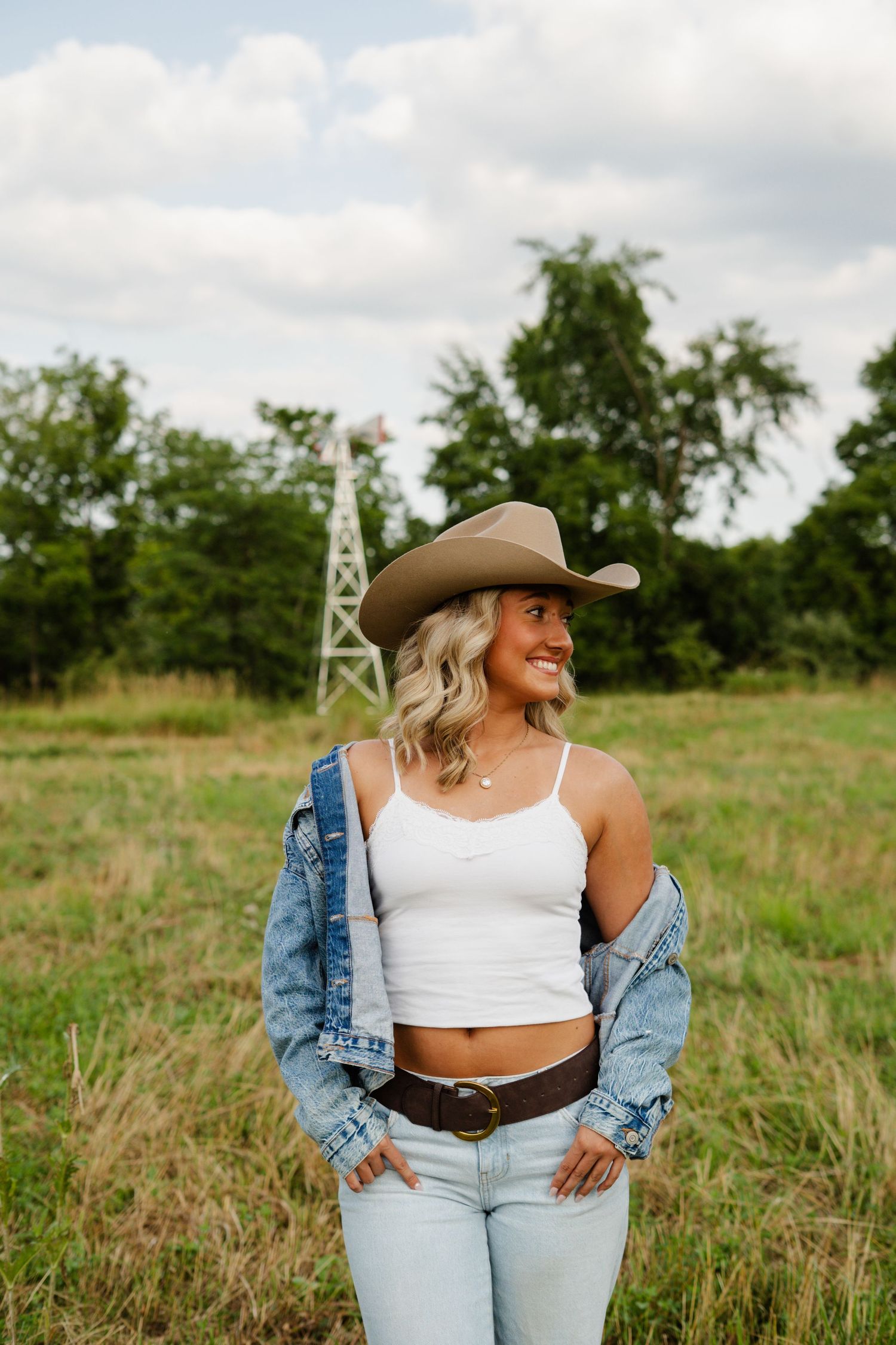 Blonde woman in white crop top, cowboy hat, and jeans smiling in a field with windmill.