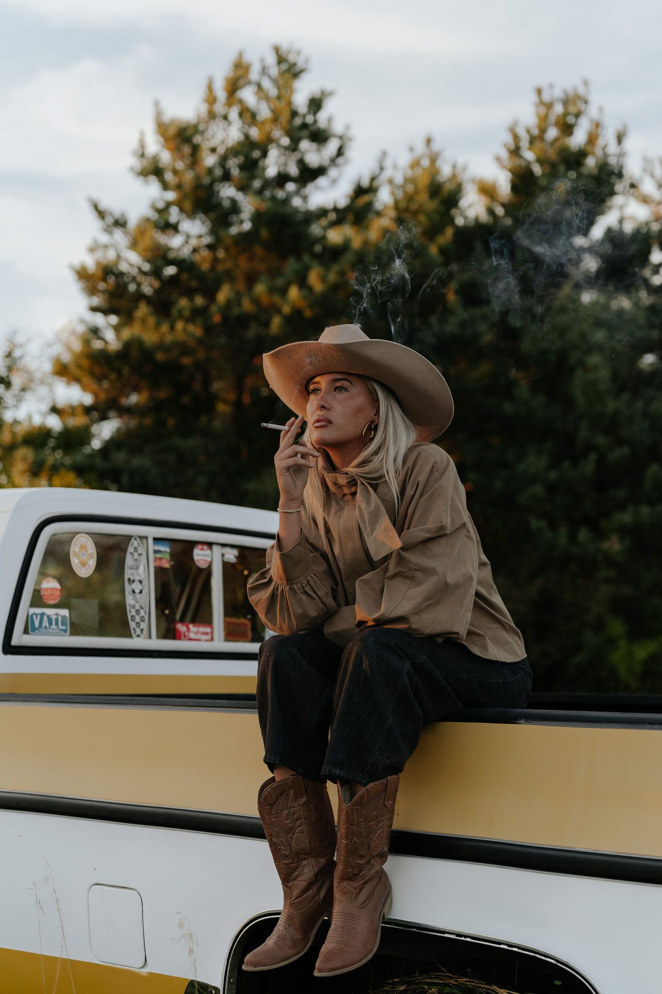 Woman in cowboy hat and boots sitting on vintage yellow truck bed smoking a cigarette outdoors.
