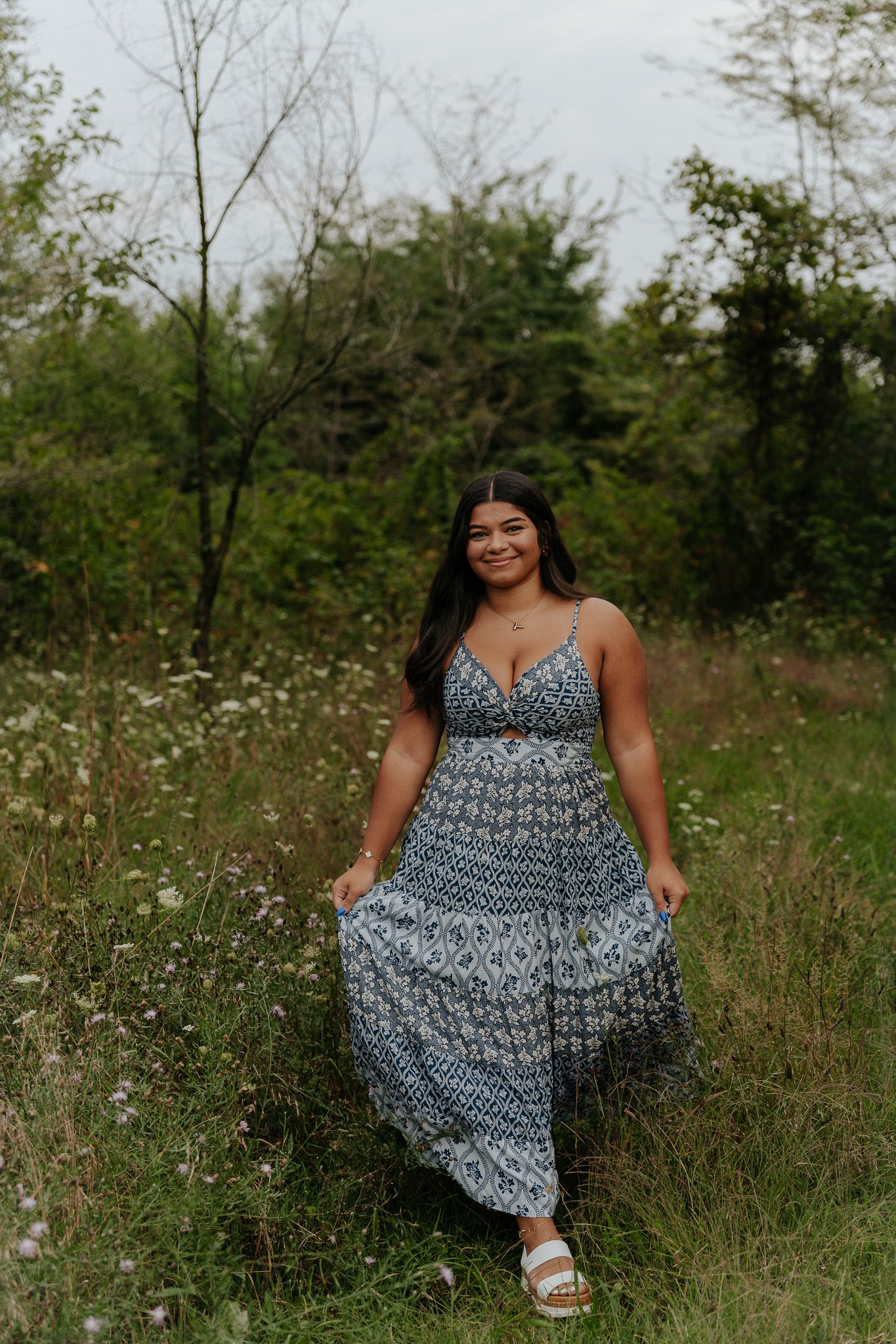 Young woman in long blue boho maxi dress walking through green meadow for senior portraits.