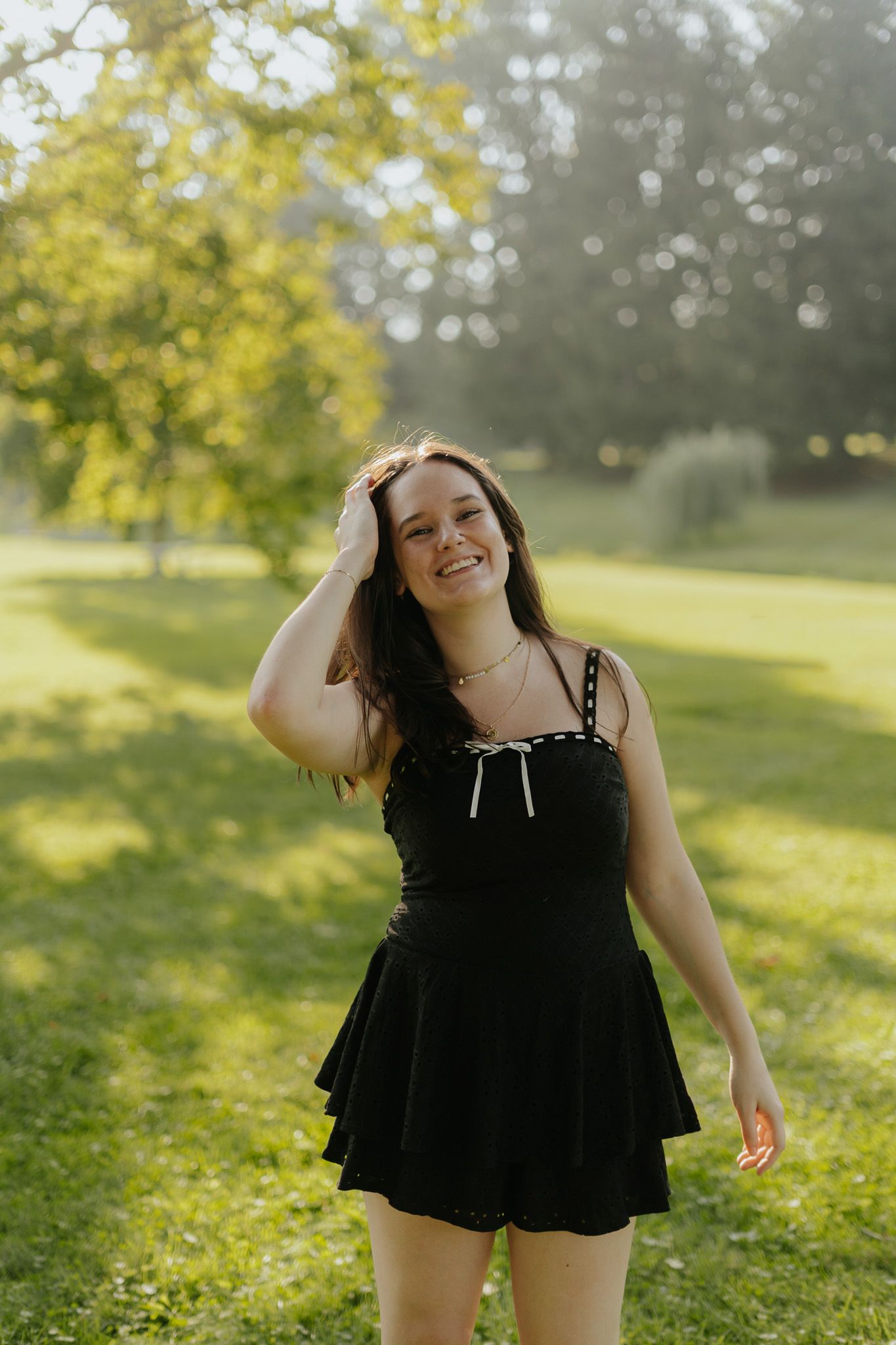 Young woman in black mini dress smiling in sunlit park.