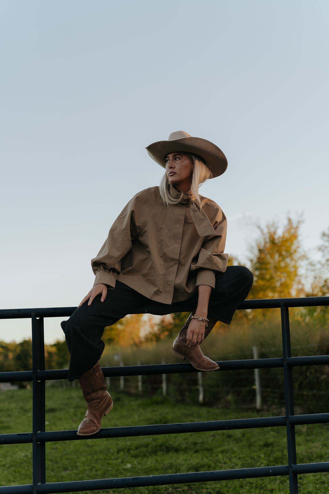 Blonde woman in cowboy hat and tan jacket sitting on metal fence at sunset.