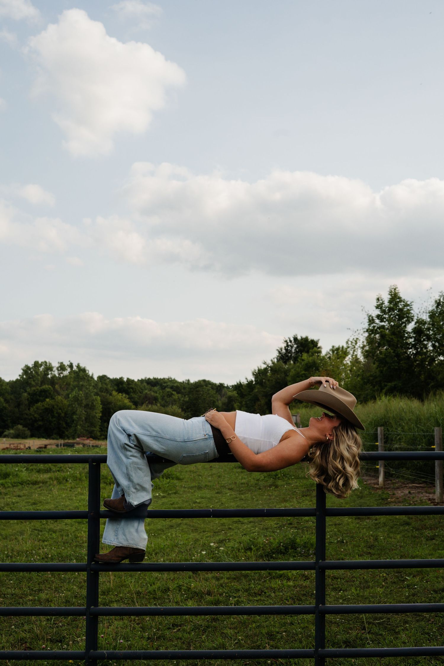 Blonde woman in cowboy hat and jeans leaning back on black farm fence in green pasture.