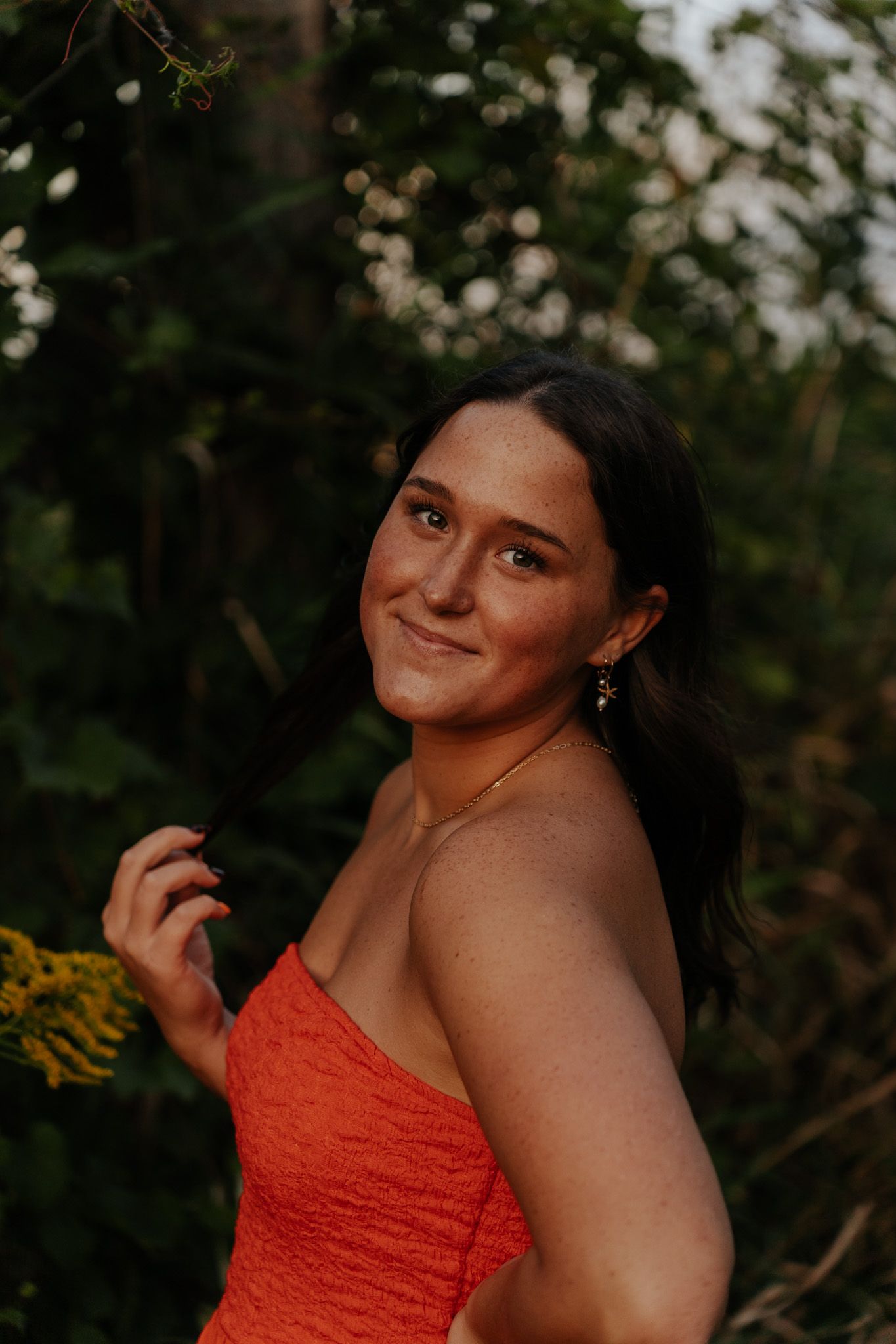 Young woman in orange strapless dress smiling outdoors among lush green foliage at golden hour.