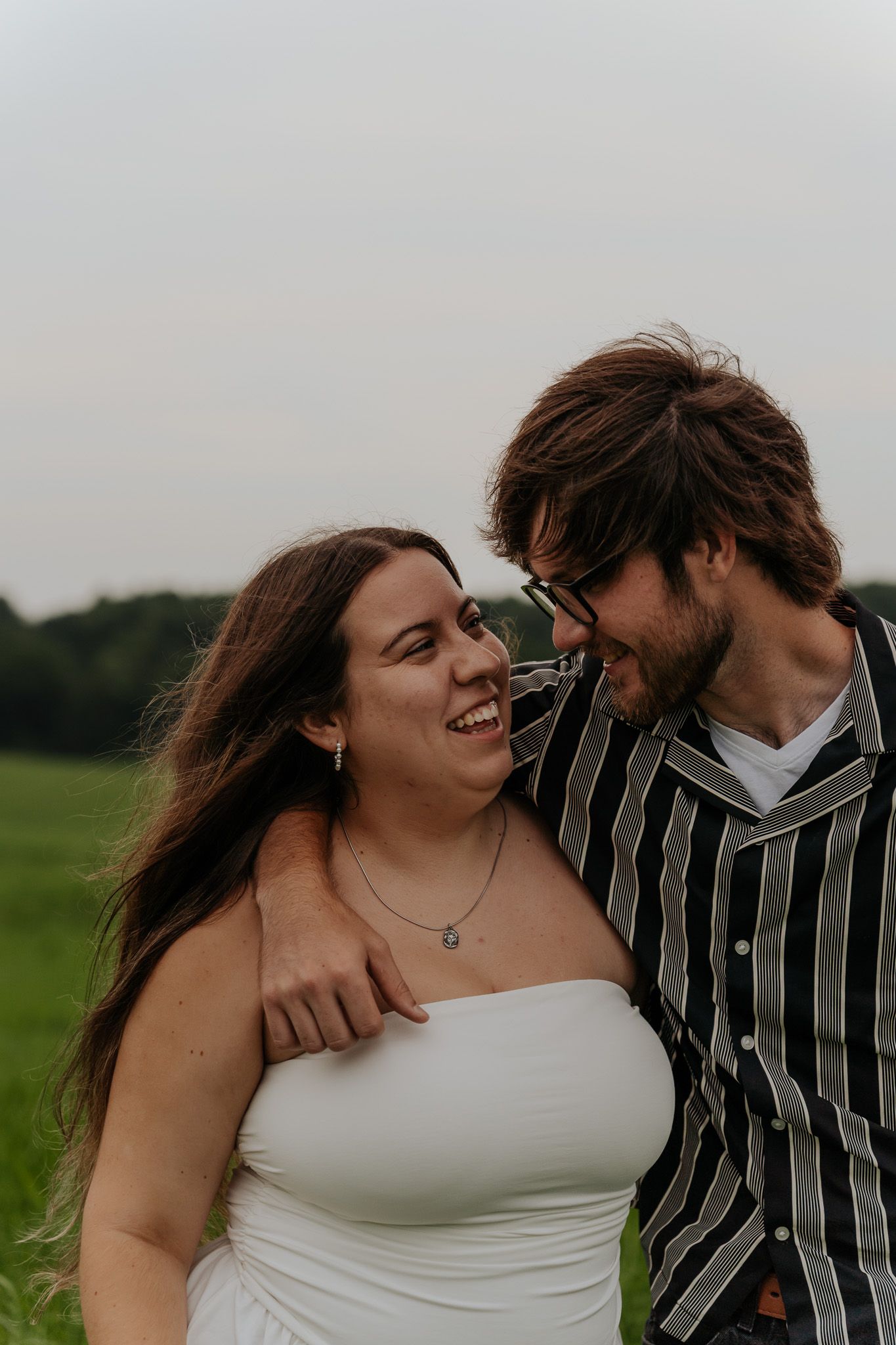 Joyful couple sharing a laugh outdoors in green field, woman in white dress, man in black striped shirt.