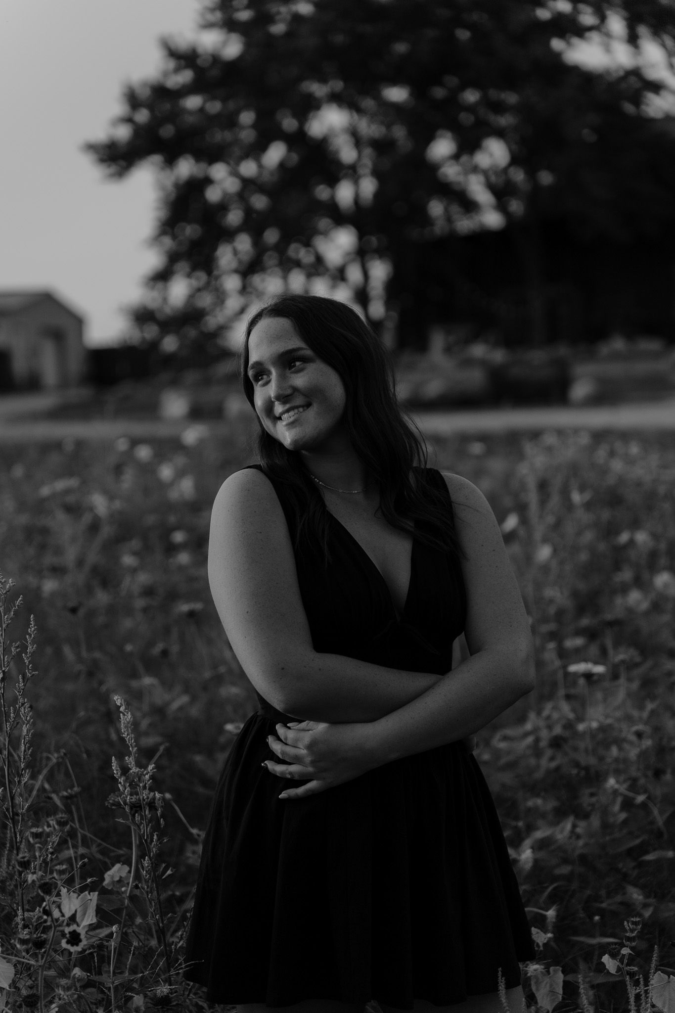 Black and white portrait of smiling young woman in black dress standing in outdoor field at sunset.
