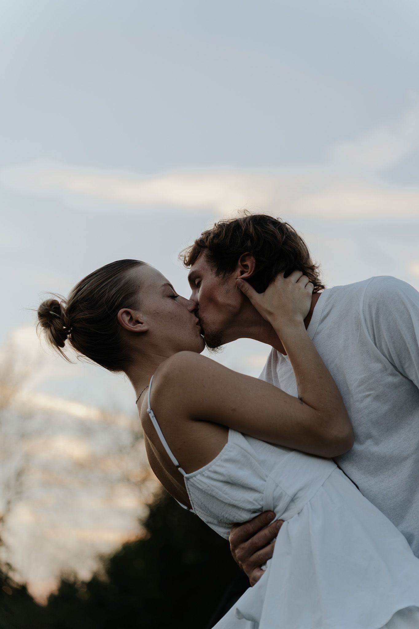 A couple shares a passionate kiss outdoors at dusk, embracing against a soft cloudy sky.