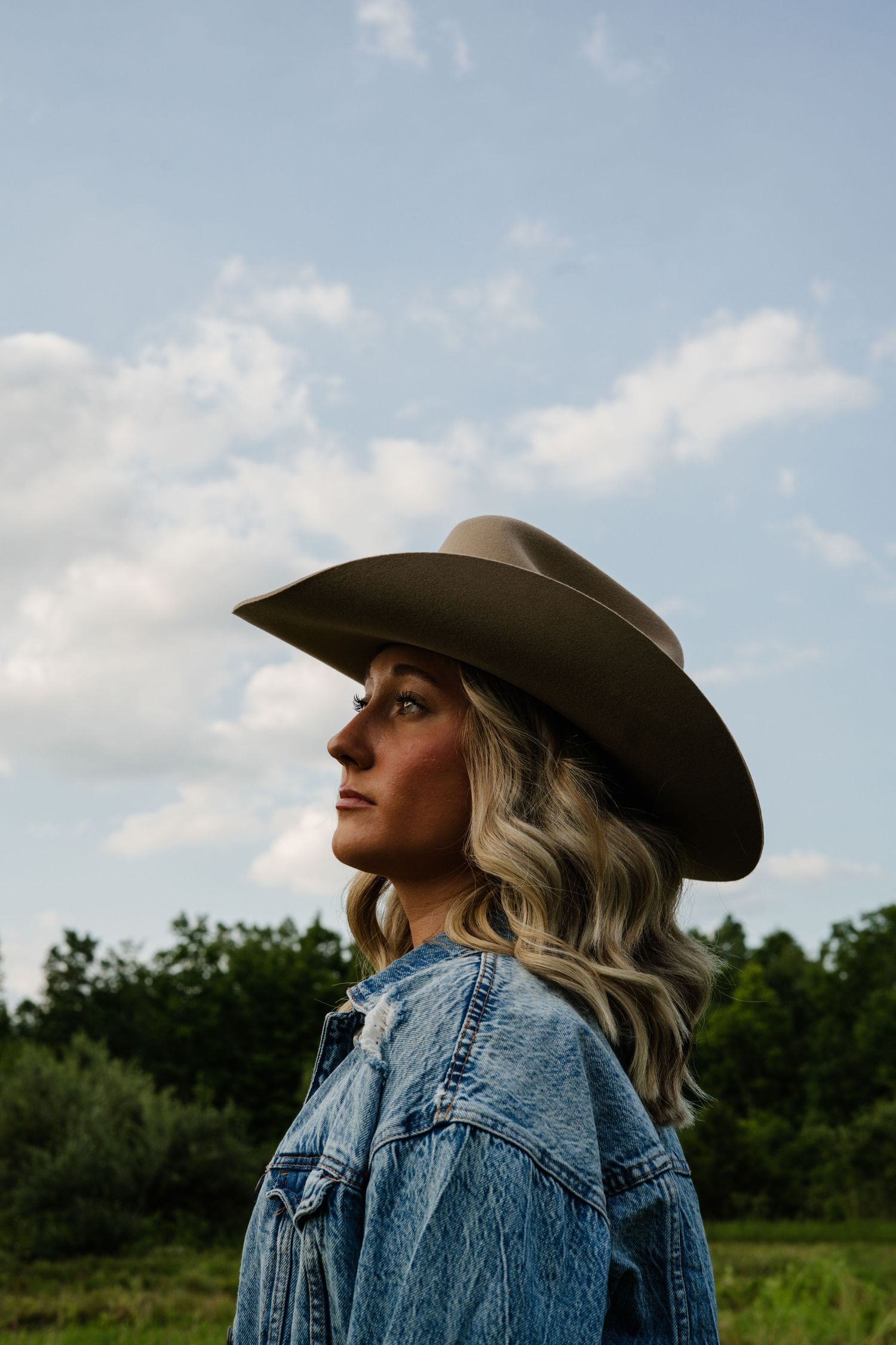 Blonde woman in denim jacket and wide-brim cowboy hat posing against cloudy sky outdoors.