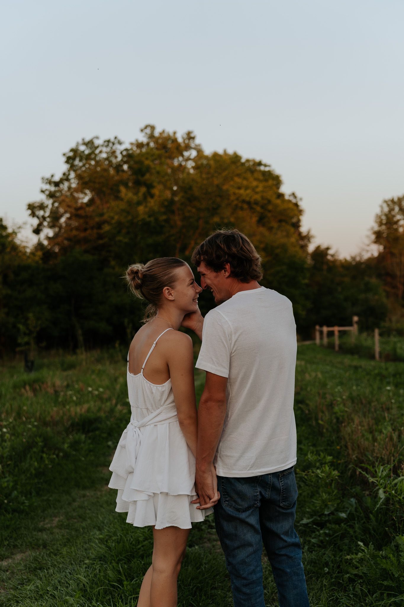 Couple in white outfits holding hands in a green field at sunset, sharing a romantic moment.
