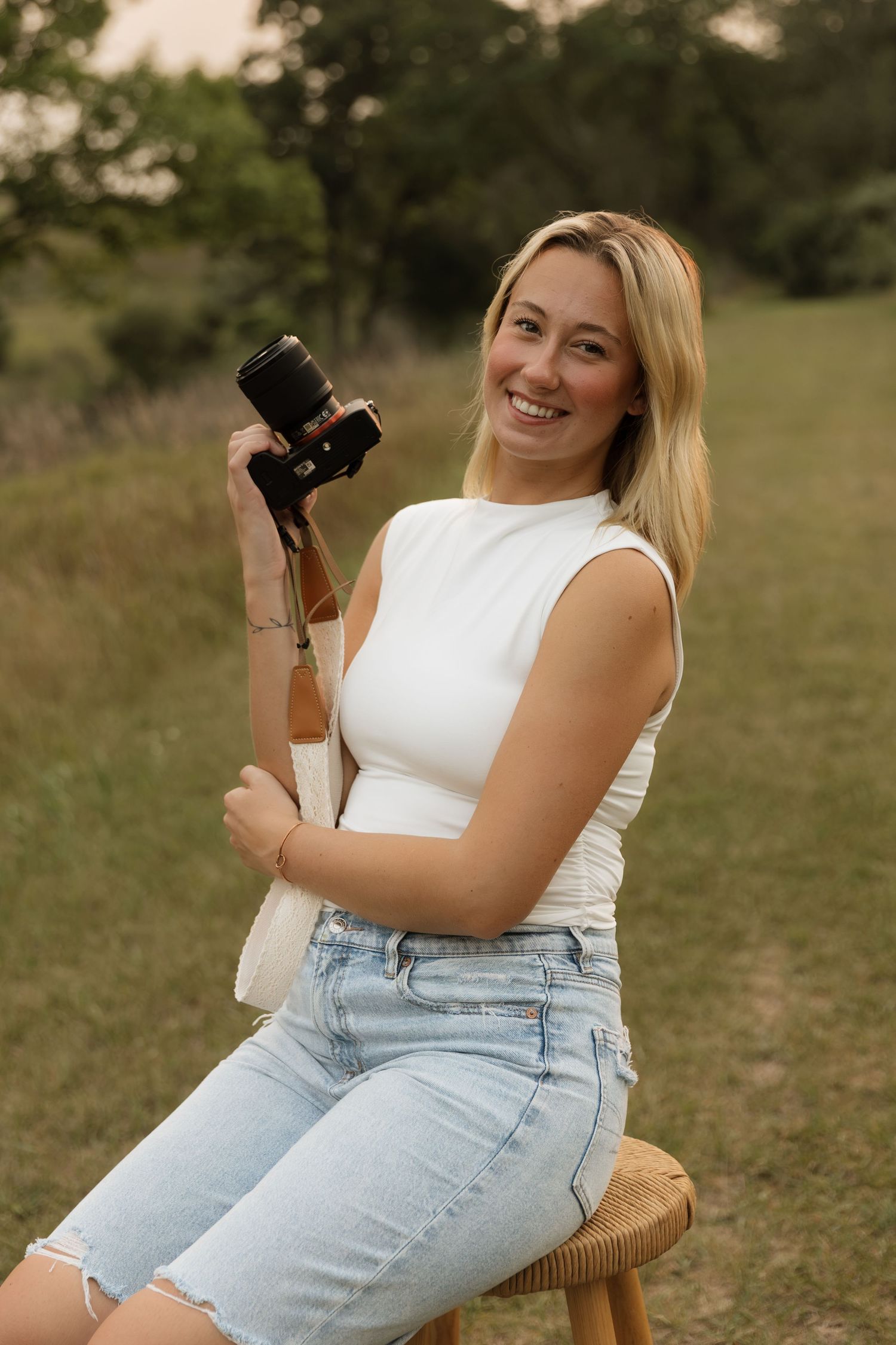 Smiling blonde woman sitting on stool holding camera outdoors in green field.