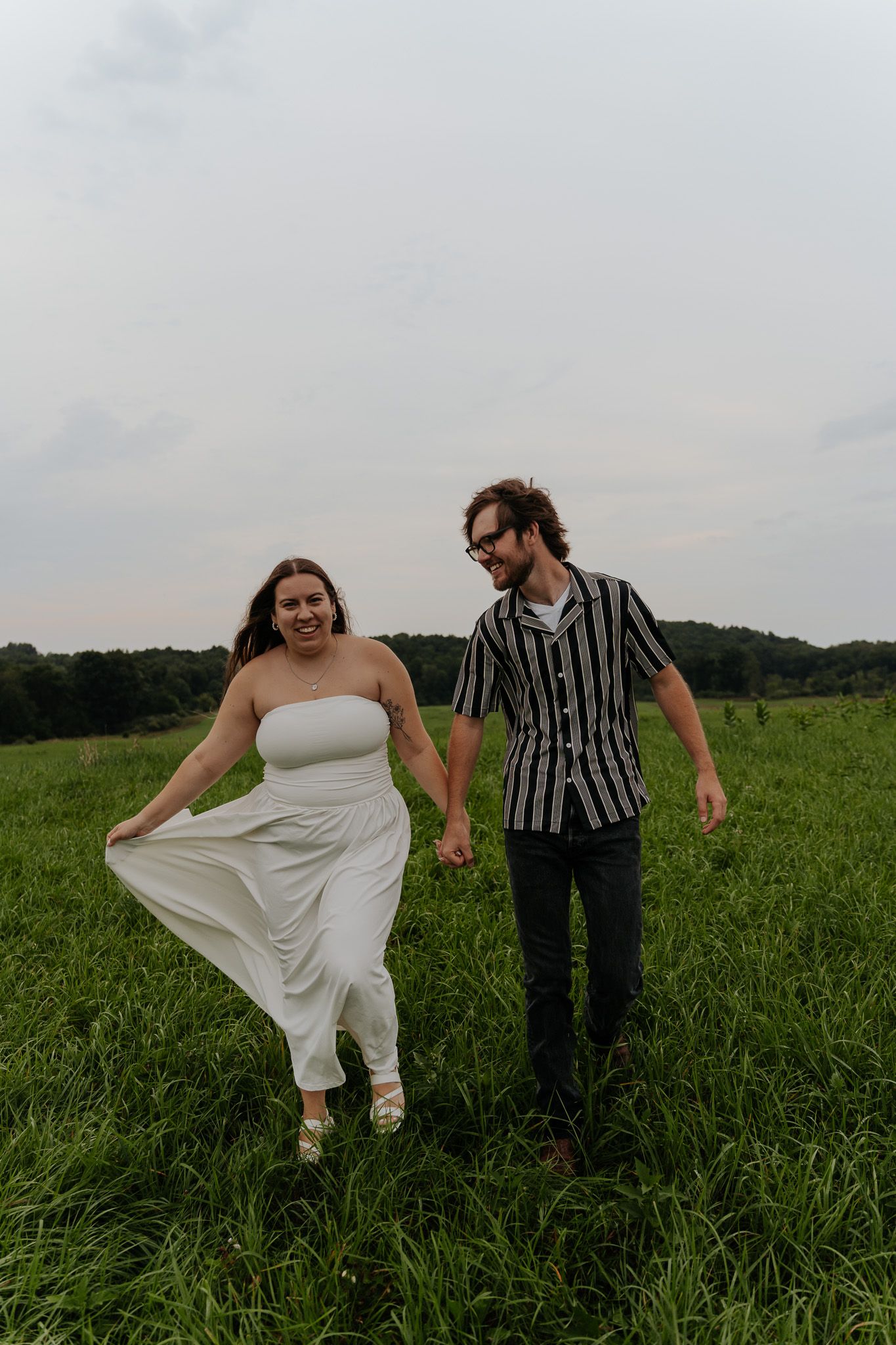 Happy couple holding hands and walking through a vibrant green field, woman in white dress lifting her skirt.
