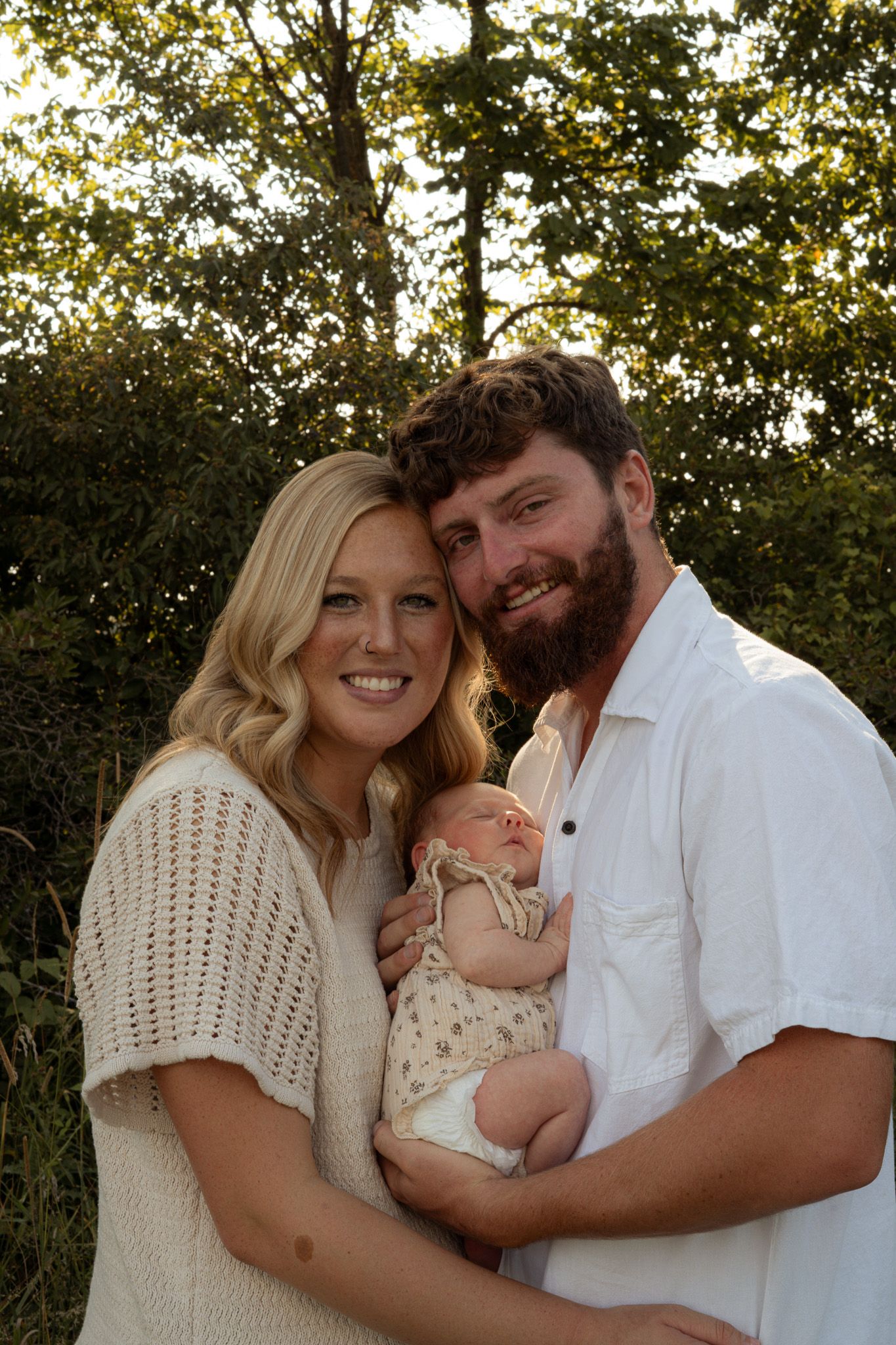Smiling young couple with blonde mother and bearded father holding newborn baby outdoors at sunset.
