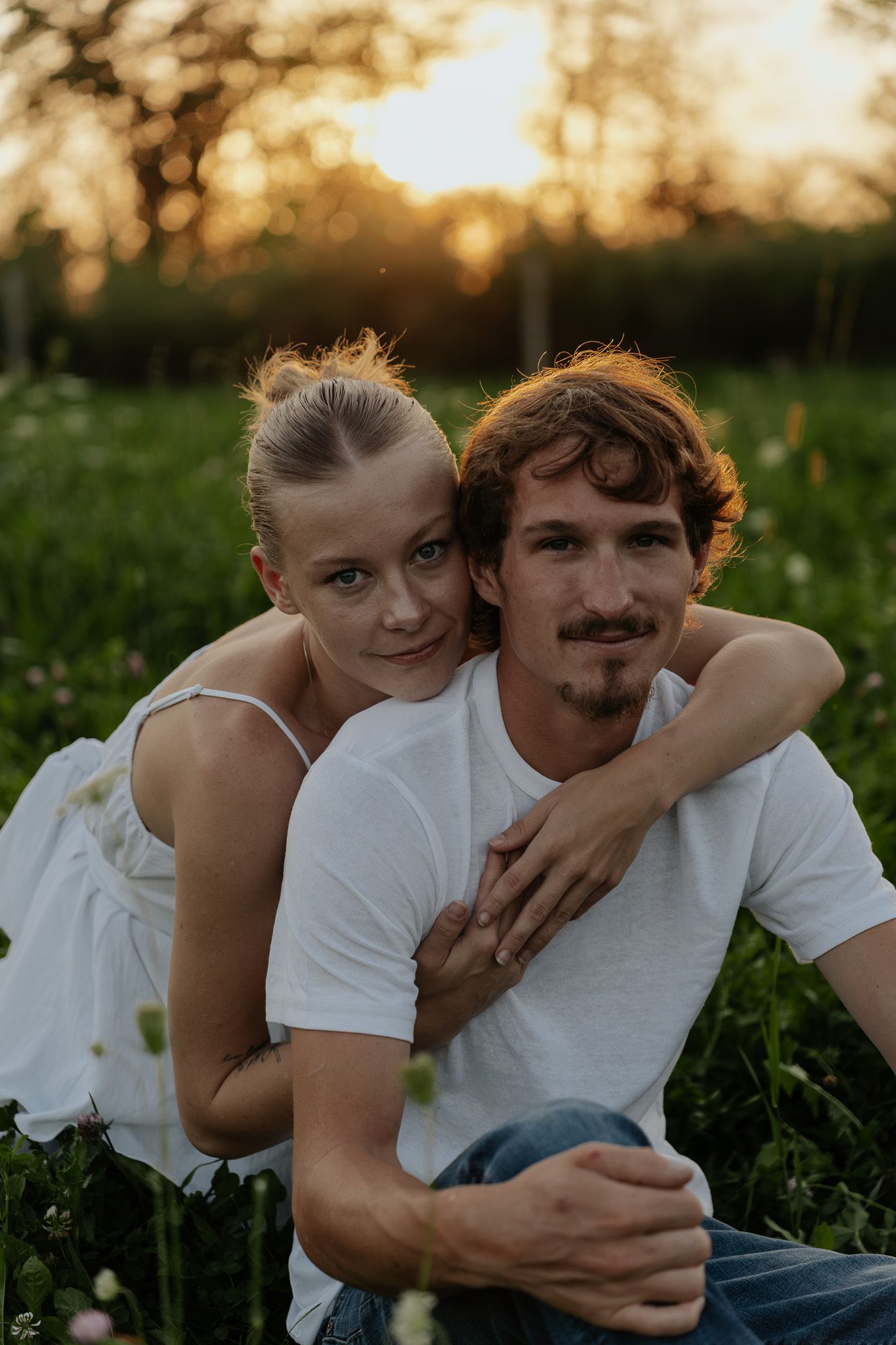 A couple embraces in a field at golden hour, woman in white dress hugging man in white tee among wildflowers.
