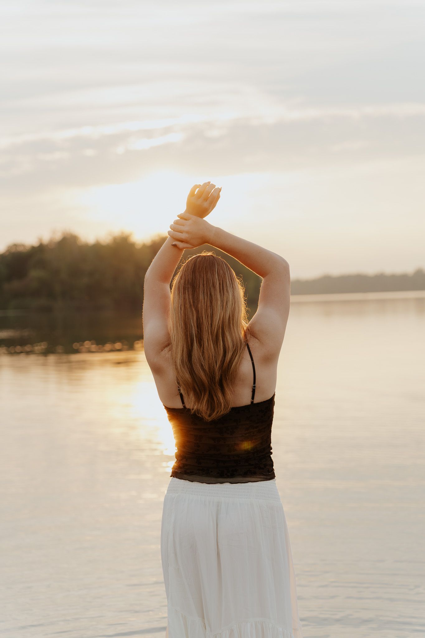 Woman in black top and white skirt raises arms toward glowing sunset over tranquil lake.