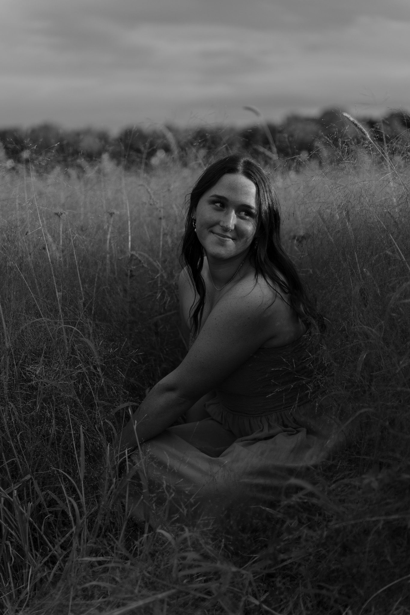 Black and white portrait of a smiling woman sitting in tall grass in a field.