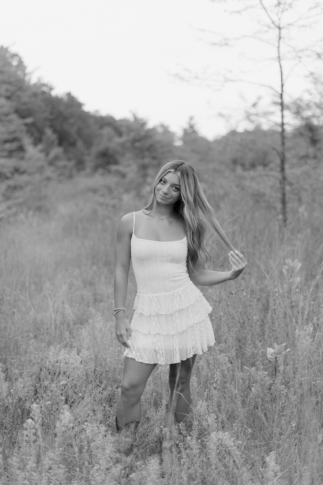 Black and white portrait of young woman in white ruffle dress standing in tall grass field.