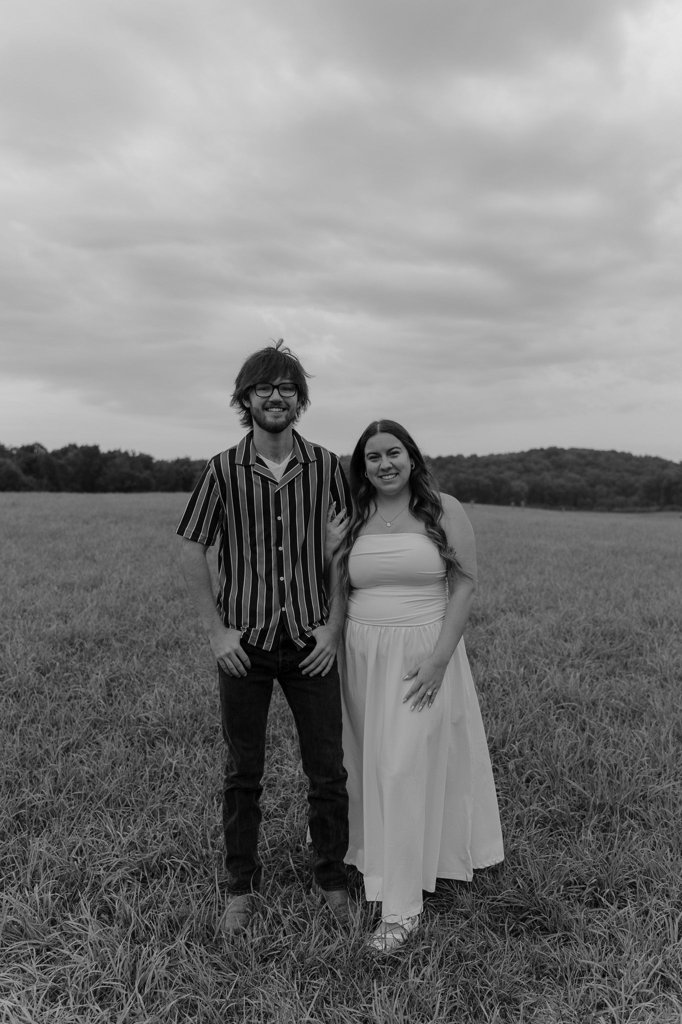 Black and white portrait of a smiling couple standing together in an open grassy field under cloudy skies.