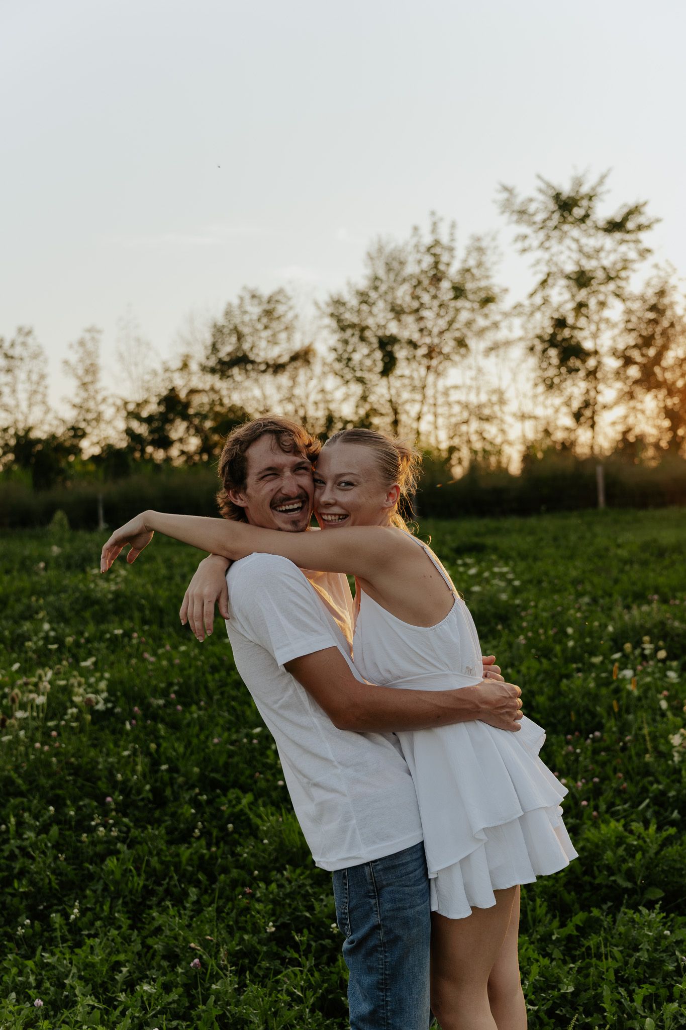 Smiling couple hugging in a lush green field during a beautiful sunset, both wearing white.