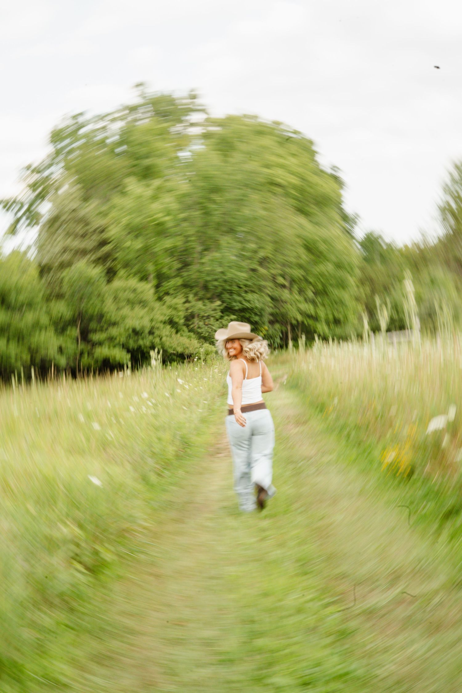 Blonde woman in cowboy hat and jeans walking through lush green meadow path with motion blur effect.