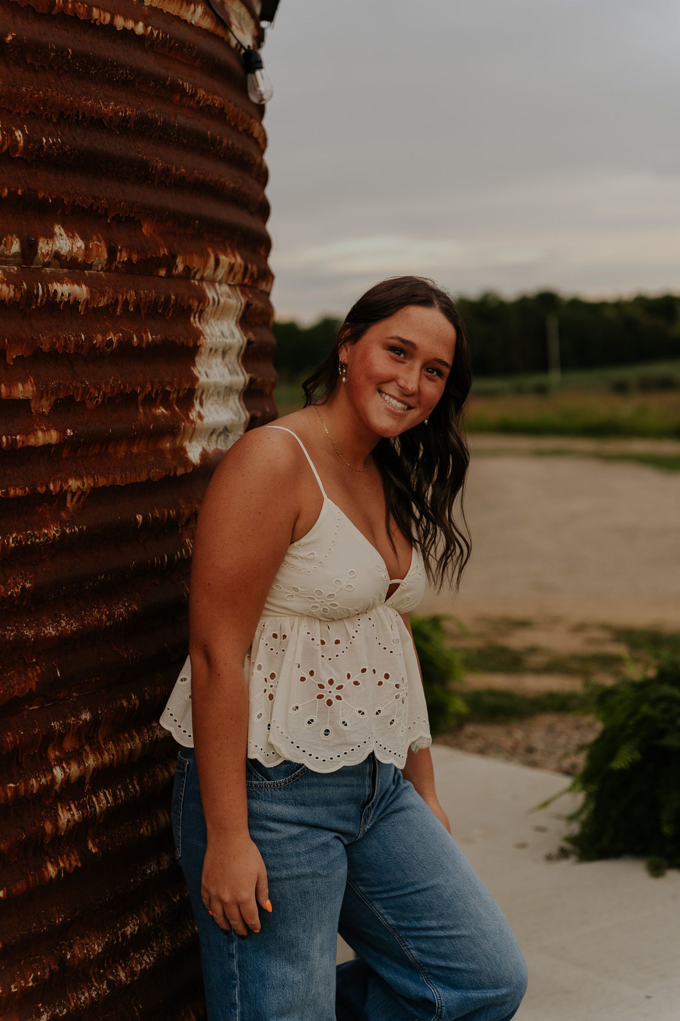 Smiling brunette woman in white eyelet top and jeans leaning against rustic metal silo.