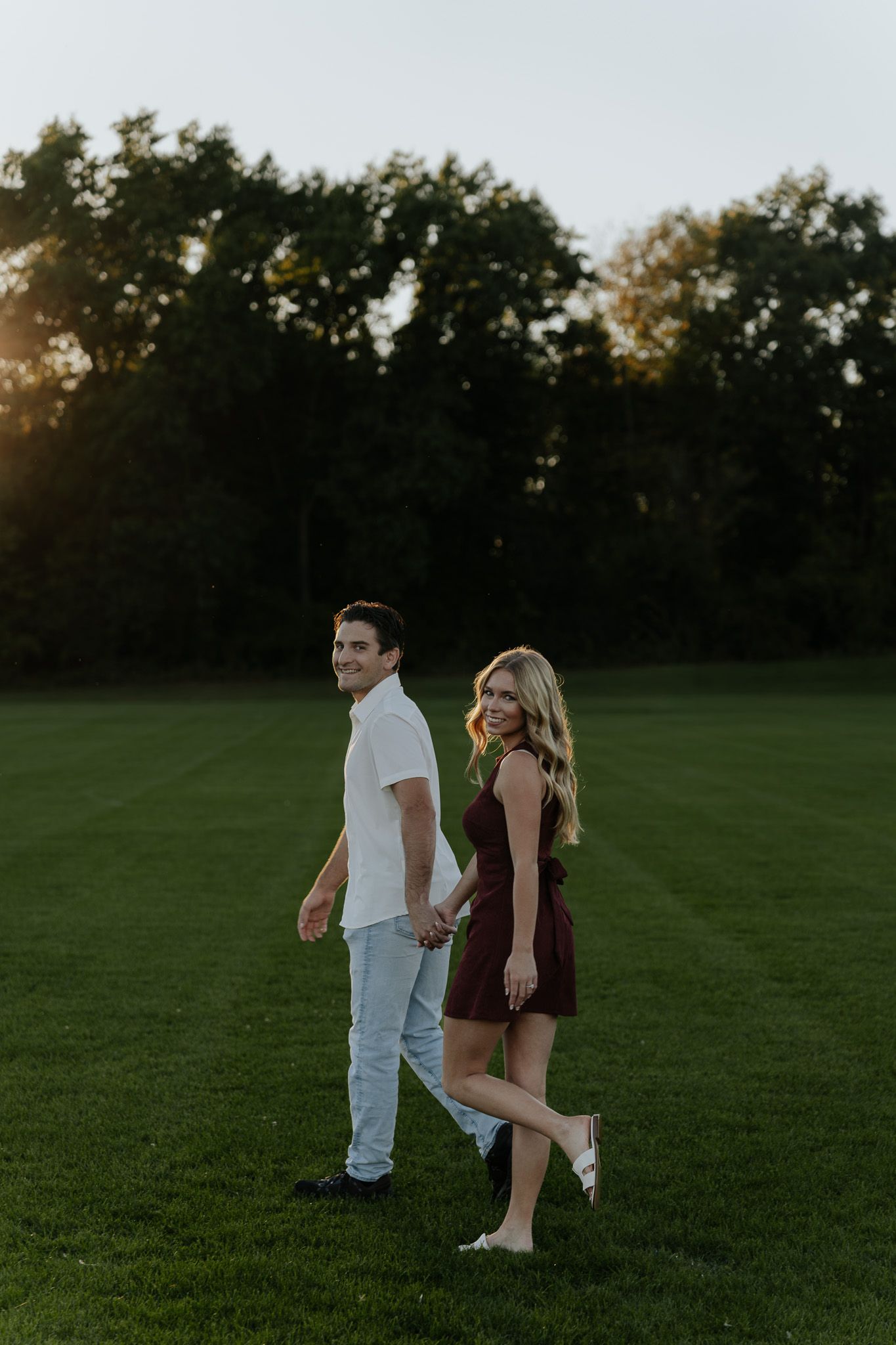 Engaged couple holding hands while walking across a green field at golden hour sunset.