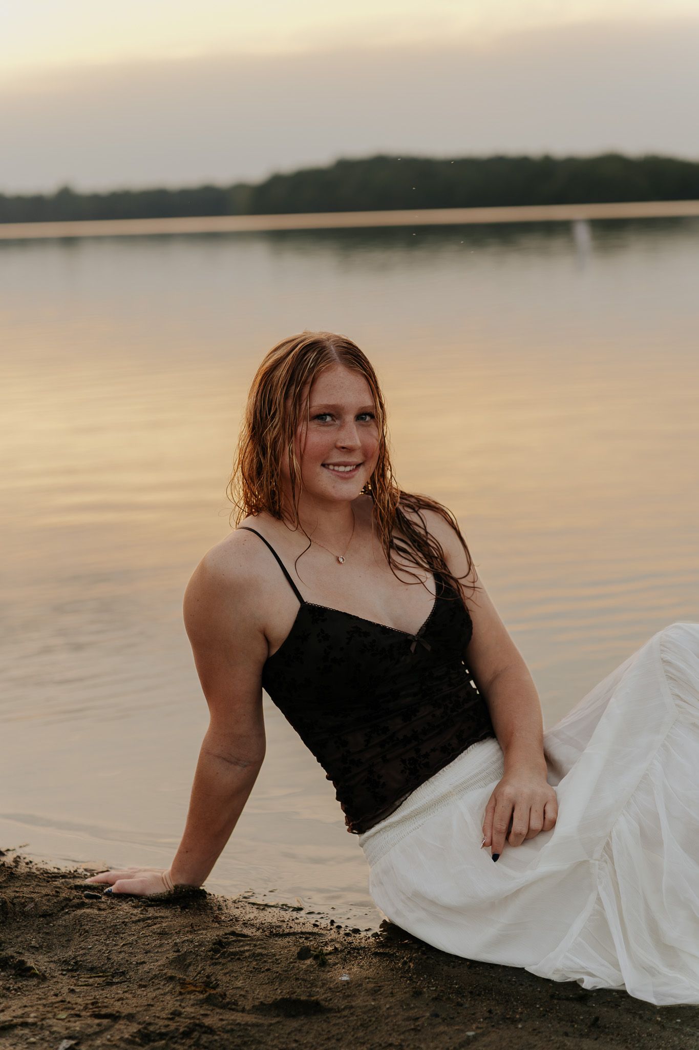 Smiling young woman with wet hair in black top and white skirt sitting on lake shore at sunset.