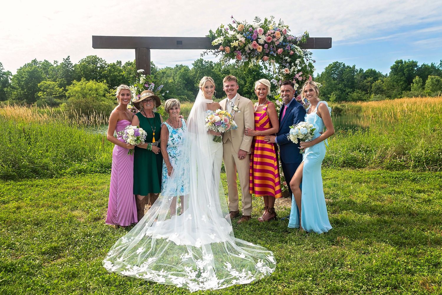 Wedding party of seven poses outdoors under a floral arch in a sunny green meadow setting.