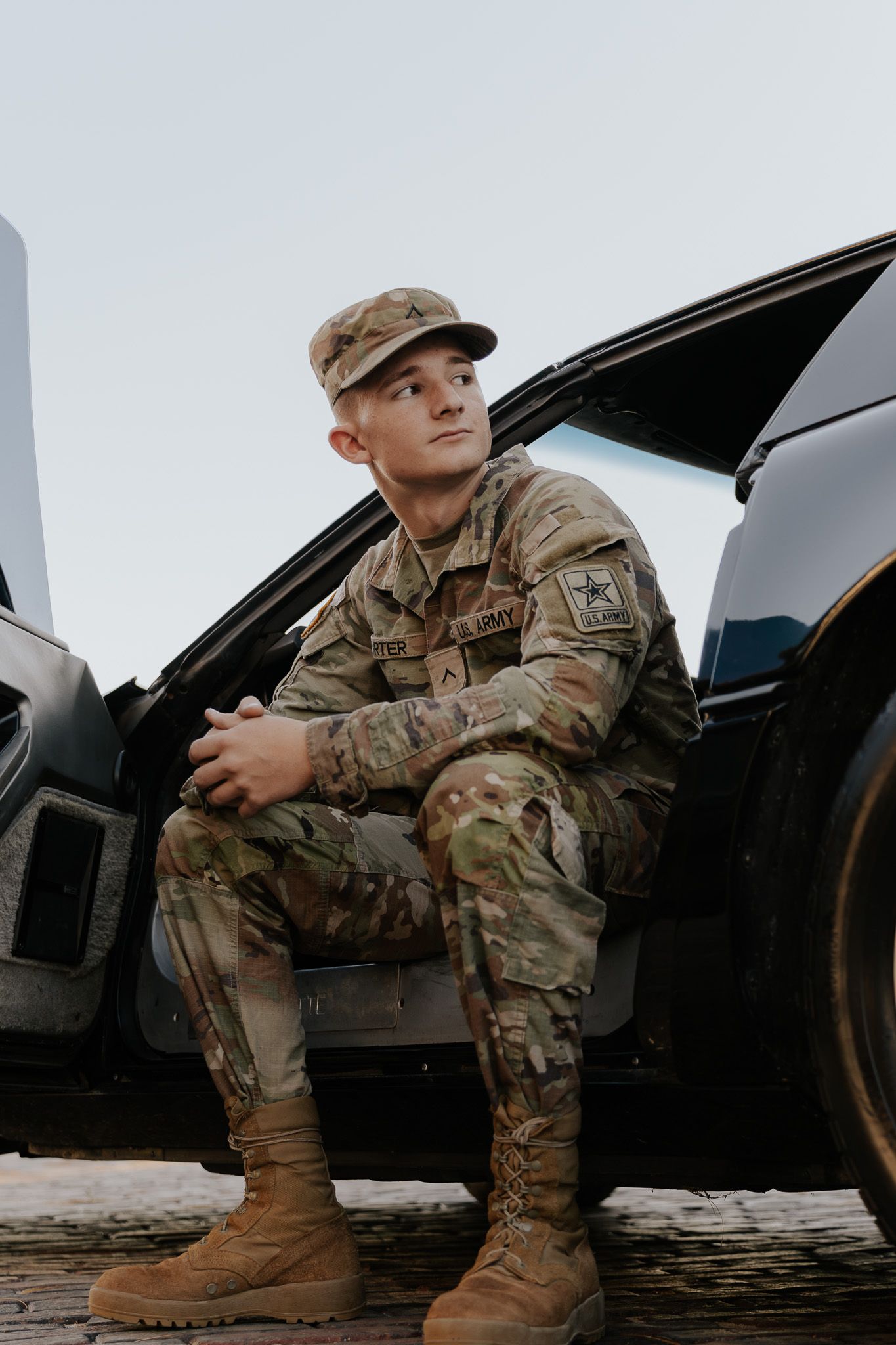 Young US Army soldier in camouflage uniform and cap sitting on vehicle door, looking thoughtfully into distance.