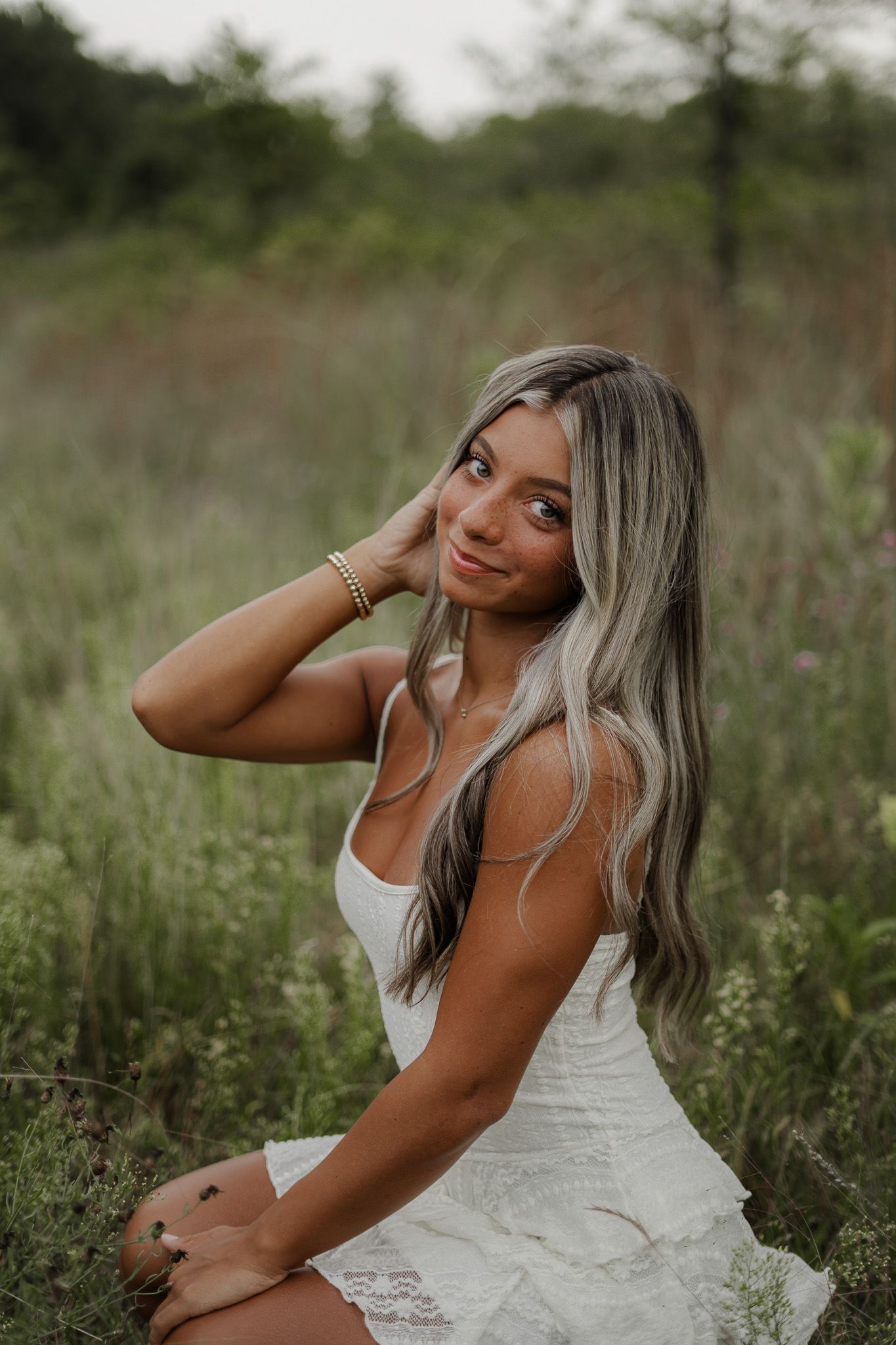 Young woman in white dress sitting in wildflower field with hand in highlighted blonde hair.