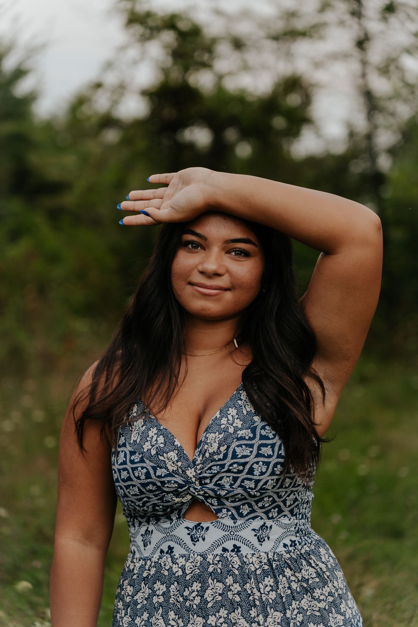 Smiling young woman in blue patterned sundress poses with raised arm in a natural outdoor green field.