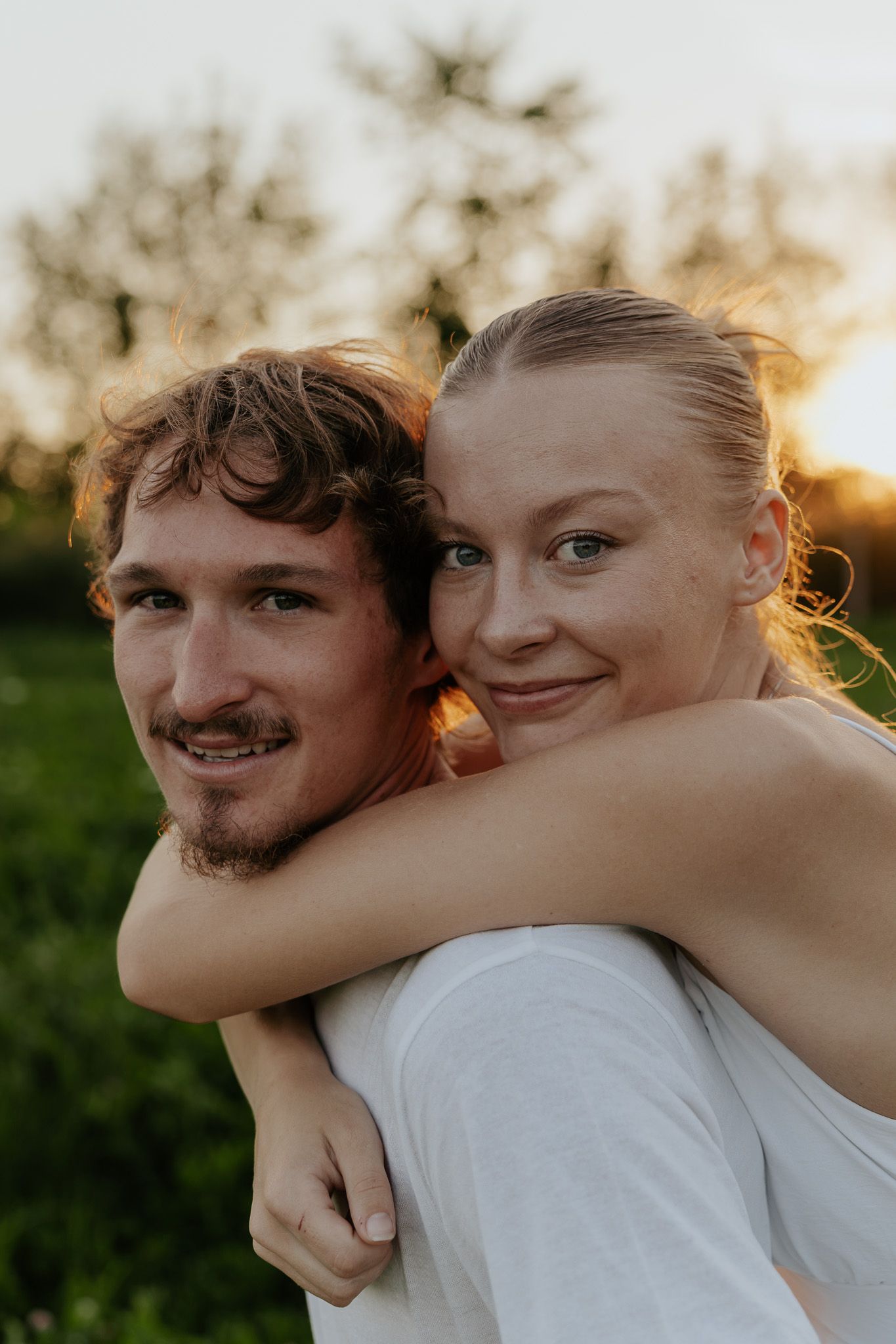 A couple embraces at golden hour, the woman hugging the man from behind while smiling outdoors.