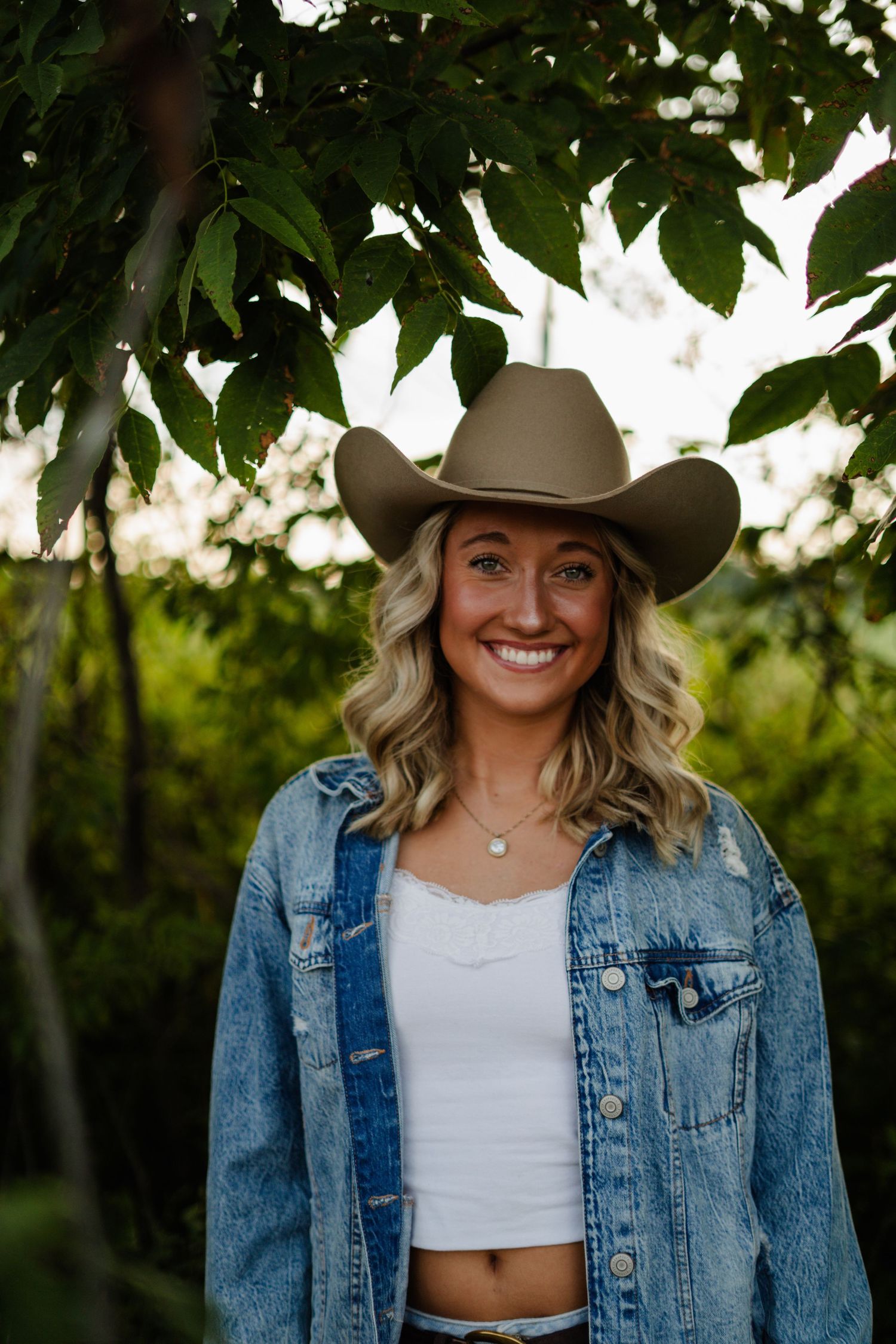 Smiling blonde woman in tan cowboy hat and denim jacket standing under green tree leaves.