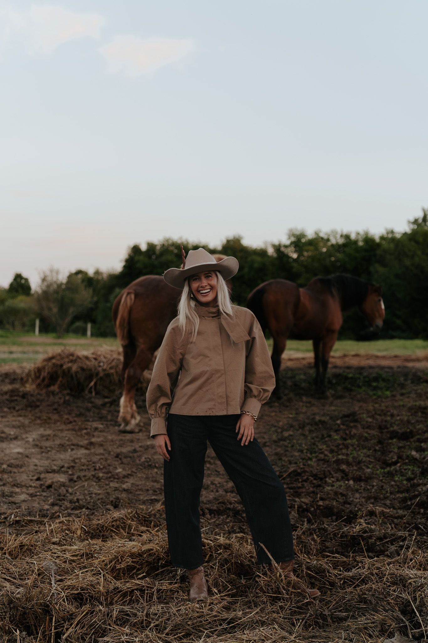 Smiling blonde woman in cowboy hat and tan jacket standing in a field with horses behind her.