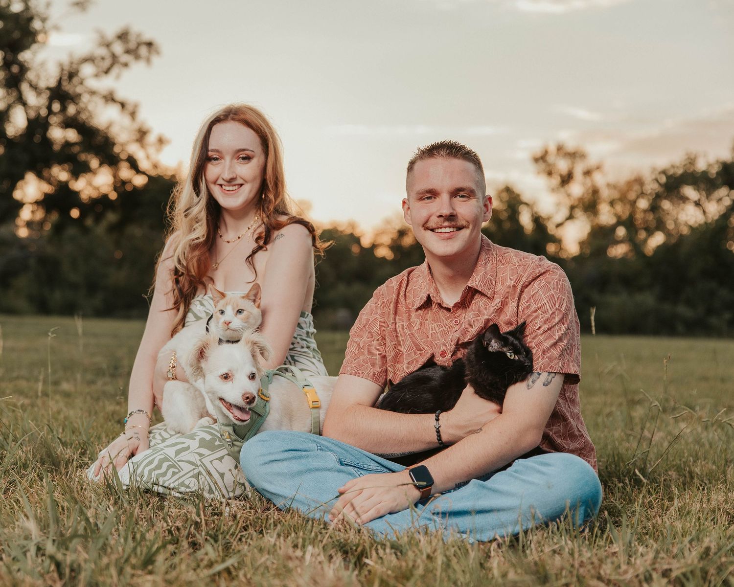 Couple sitting on grass at sunset holding small dogs, smiling outdoors in a park setting.