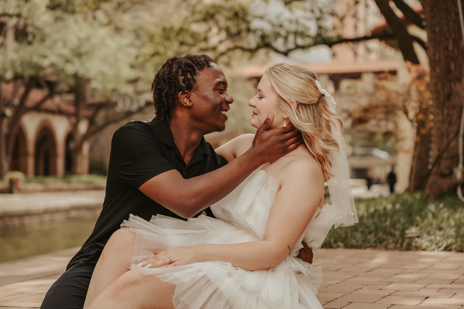 Couple in wedding attire sharing a romantic moment outdoors, groom in black shirt and bride in white dress smiling at each other.