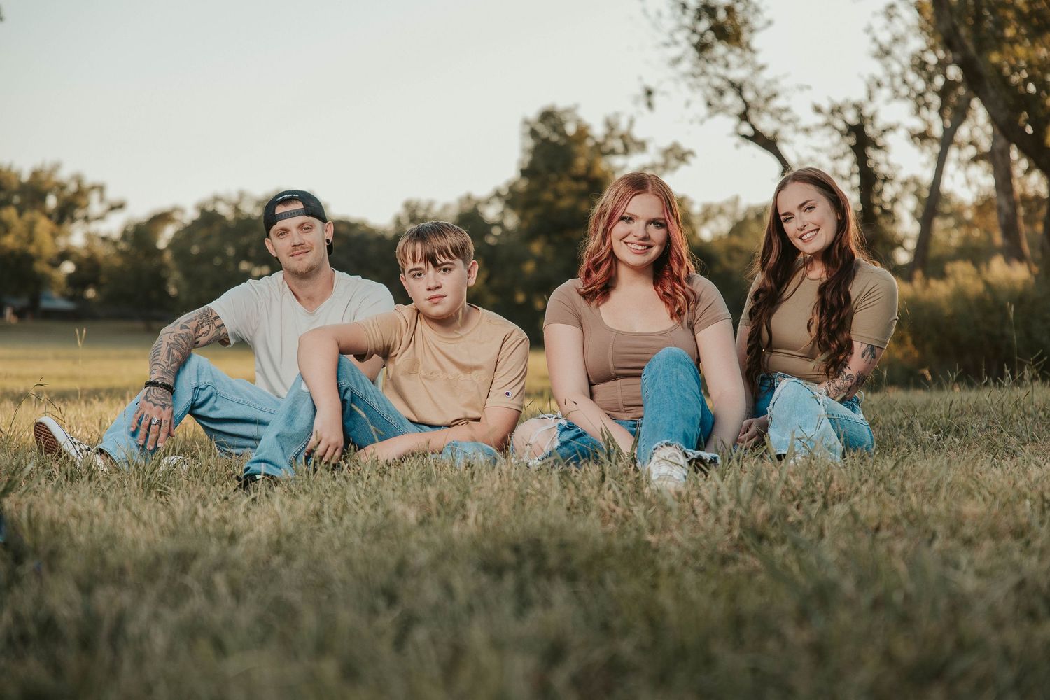 Four young adults sitting on grass in a park, posing for a casual outdoor group photo at golden hour.