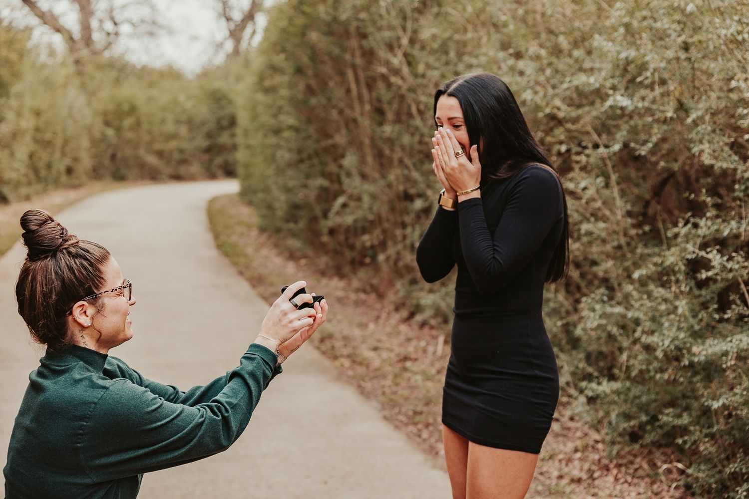 Man kneeling on a path proposing to a surprised woman covering her face, surrounded by lush greenery.