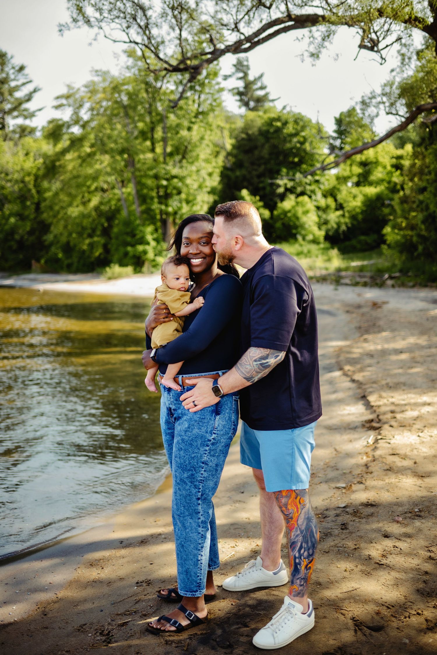 Mom holding son while dad kisses mom on the cheek while on the beach in summer. Ottawa Family Photography.