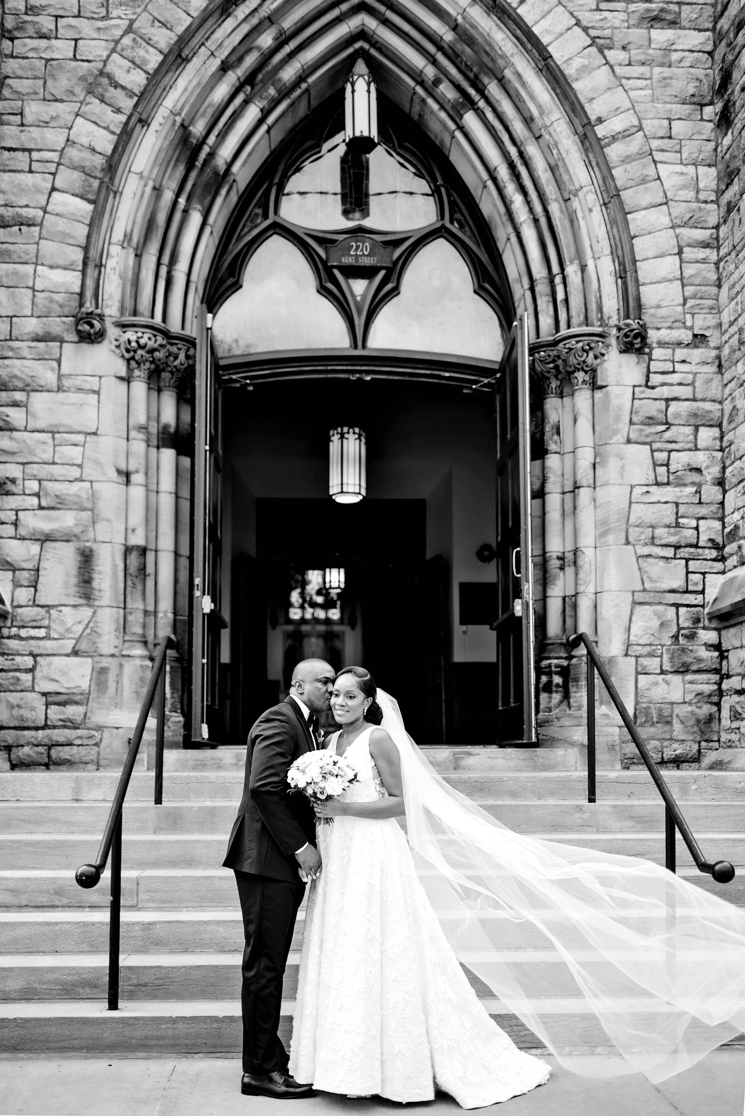 Groom kissing brides cheek outside church steps with veil blowing in the wind. Ottawa Wedding Photography. Ottawa Wedding Videography.